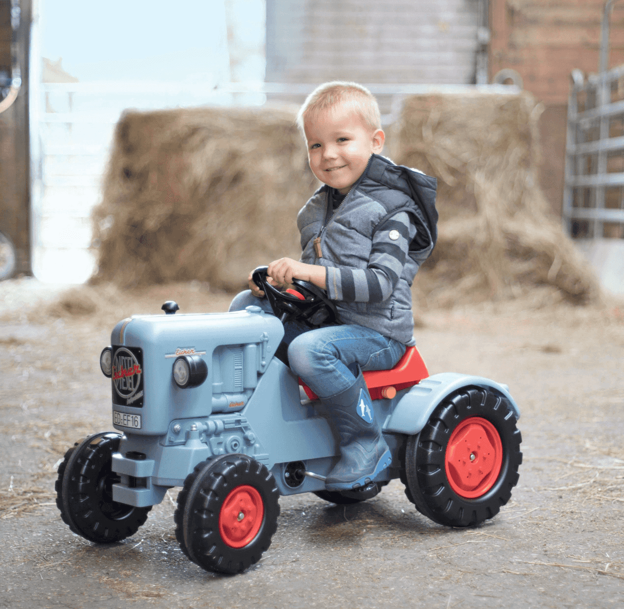 Un niño pequeño sonríe mientras monta en un tractor de juguete azul dentro de un granero.