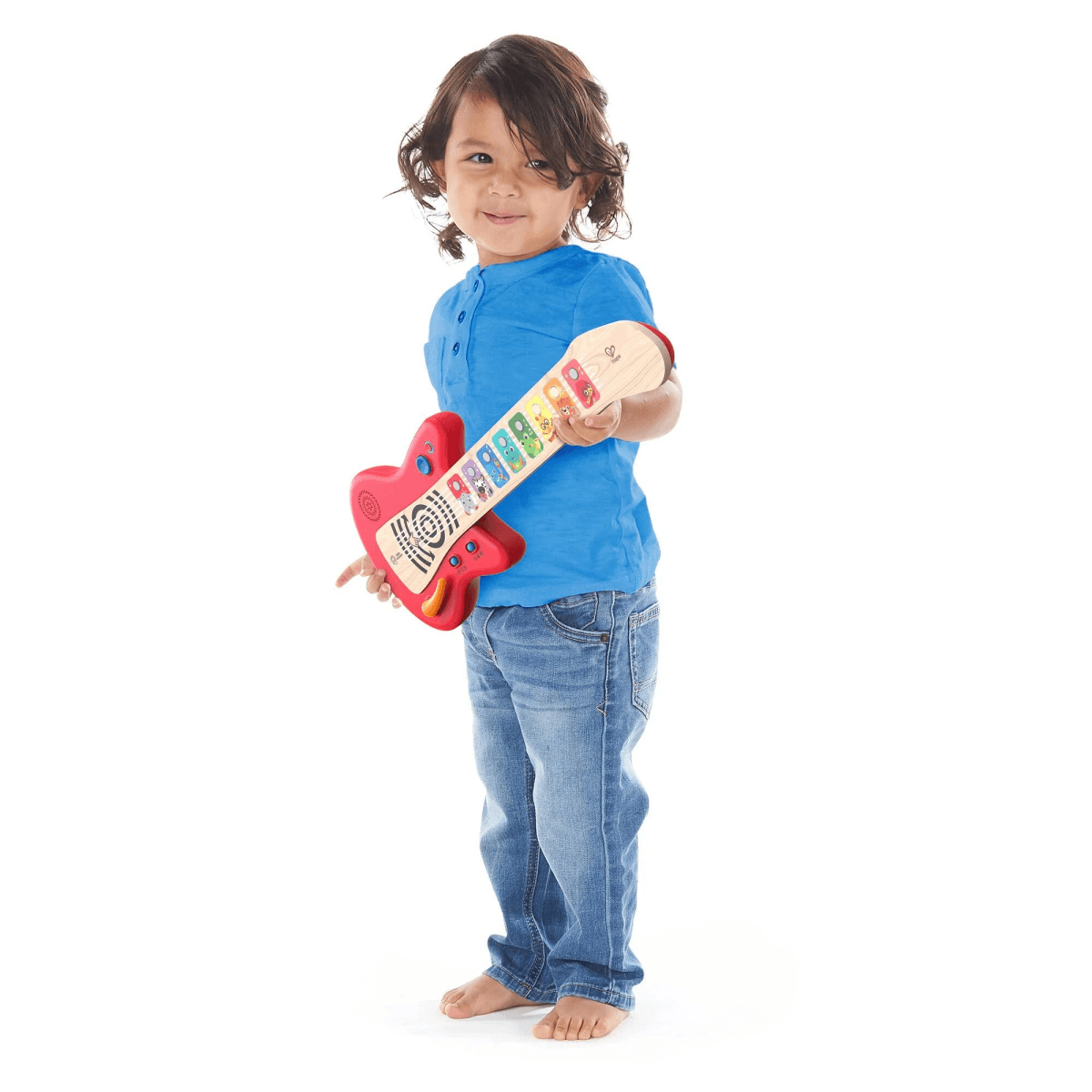 Un niño pequeño sonriente con camisa azul sostiene una guitarra de juguete roja sobre fondo blanco.