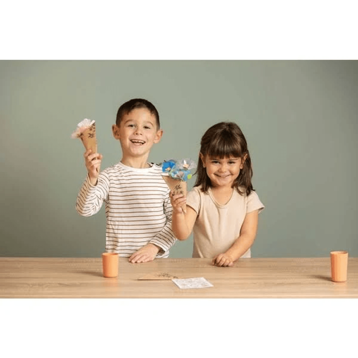 Un niño y una niña sonrientes detrás de una mesa de madera sostienen conos de flores.