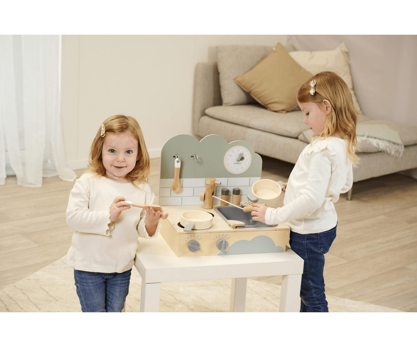 Zwei kleine Mädchen spielen in einem hellen Wohnzimmer mit einer kleinen Holzküche auf einem Tisch.