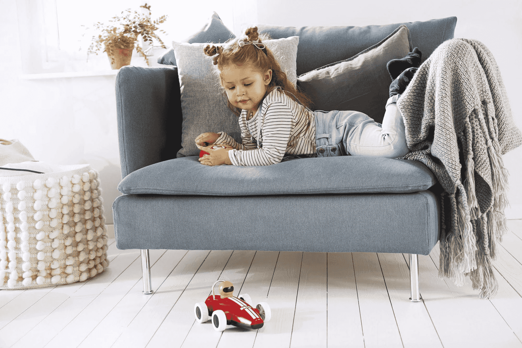 Ein Kind liegt auf einem grauen Sofa und spielt mit einem kleinen roten Spielzeugauto auf dem weißen Holzboden.