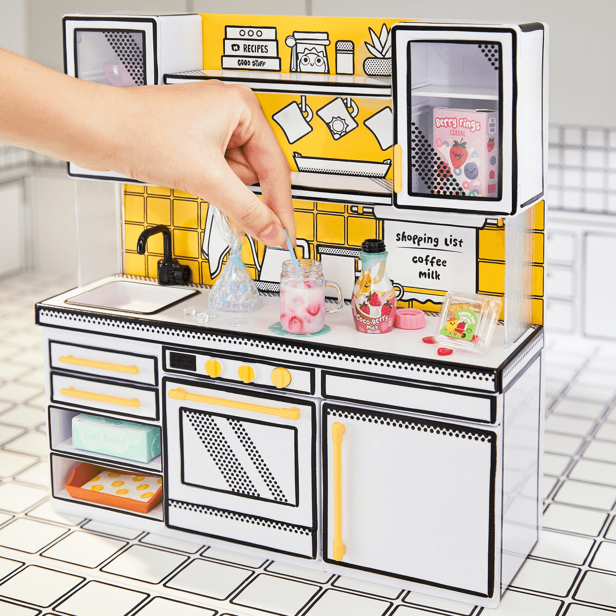 Una mano revuelve una bebida rosa en una cocina de juguete en miniatura con detalles amarillos y fregadero.