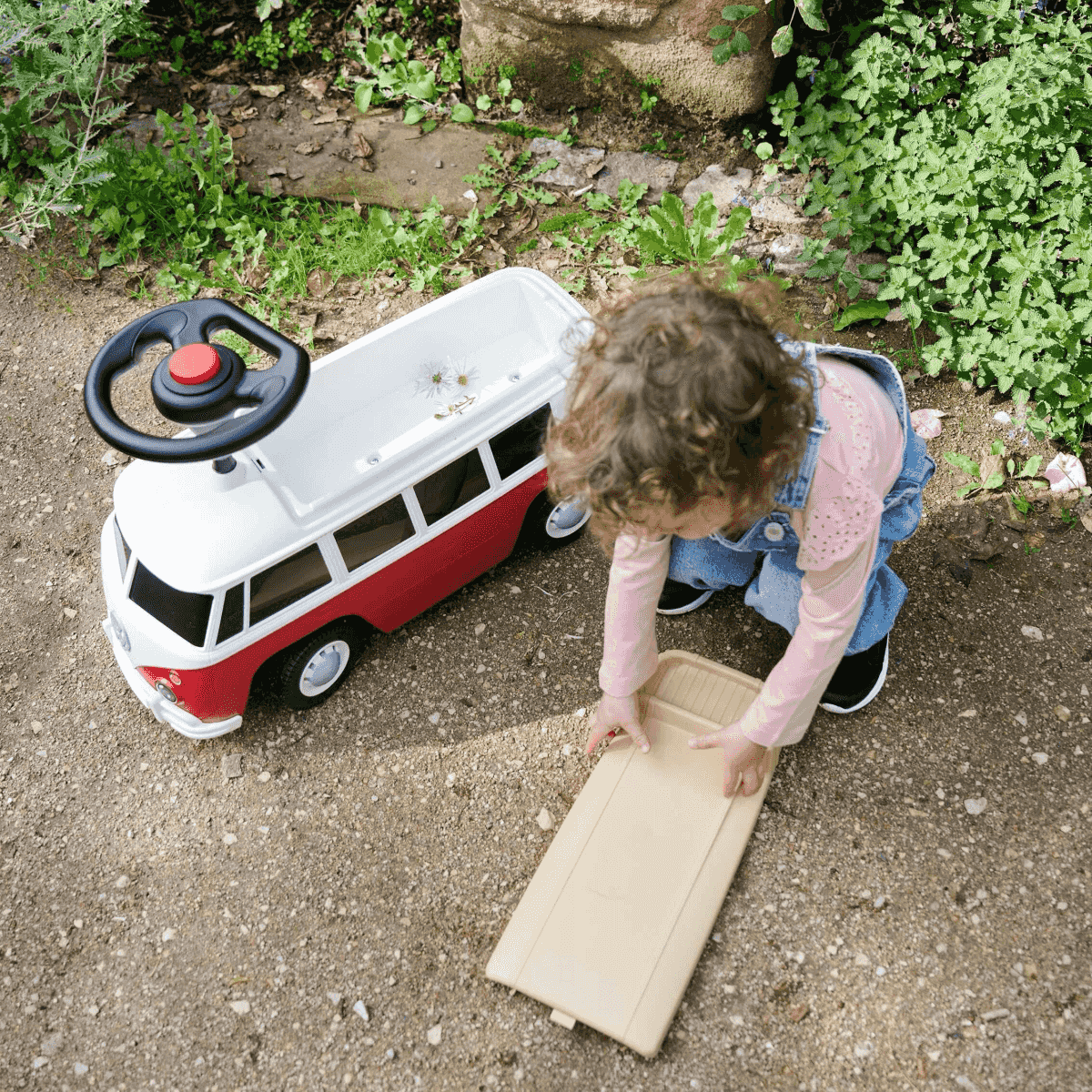 Kleinkind kniet im Garten neben einem rot-weißen Spielzeugbus und hält ein Brett.