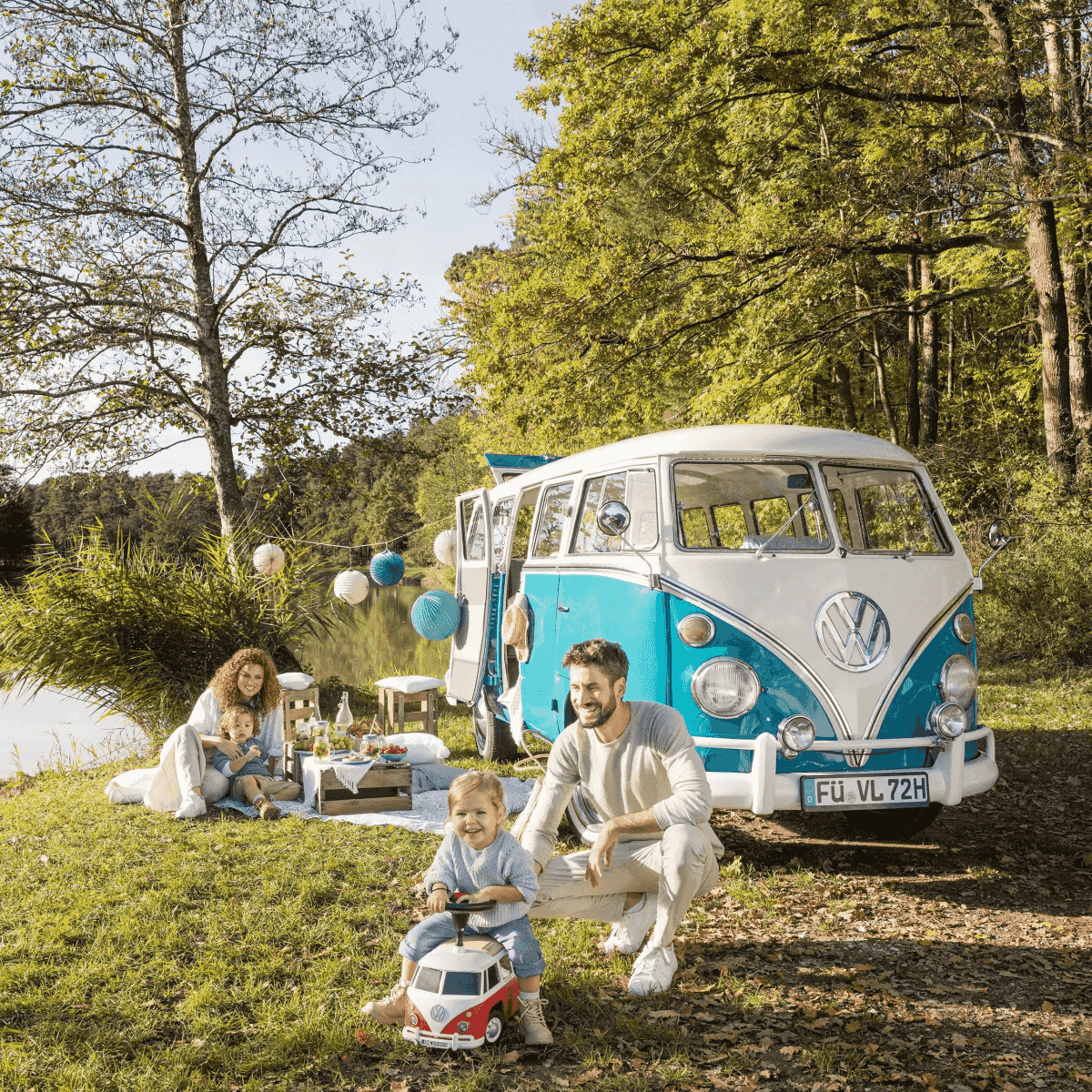 Eine Familie mit zwei Kindern macht ein Picknick am See neben einem klassischen blauen und weißen VW-Bus.