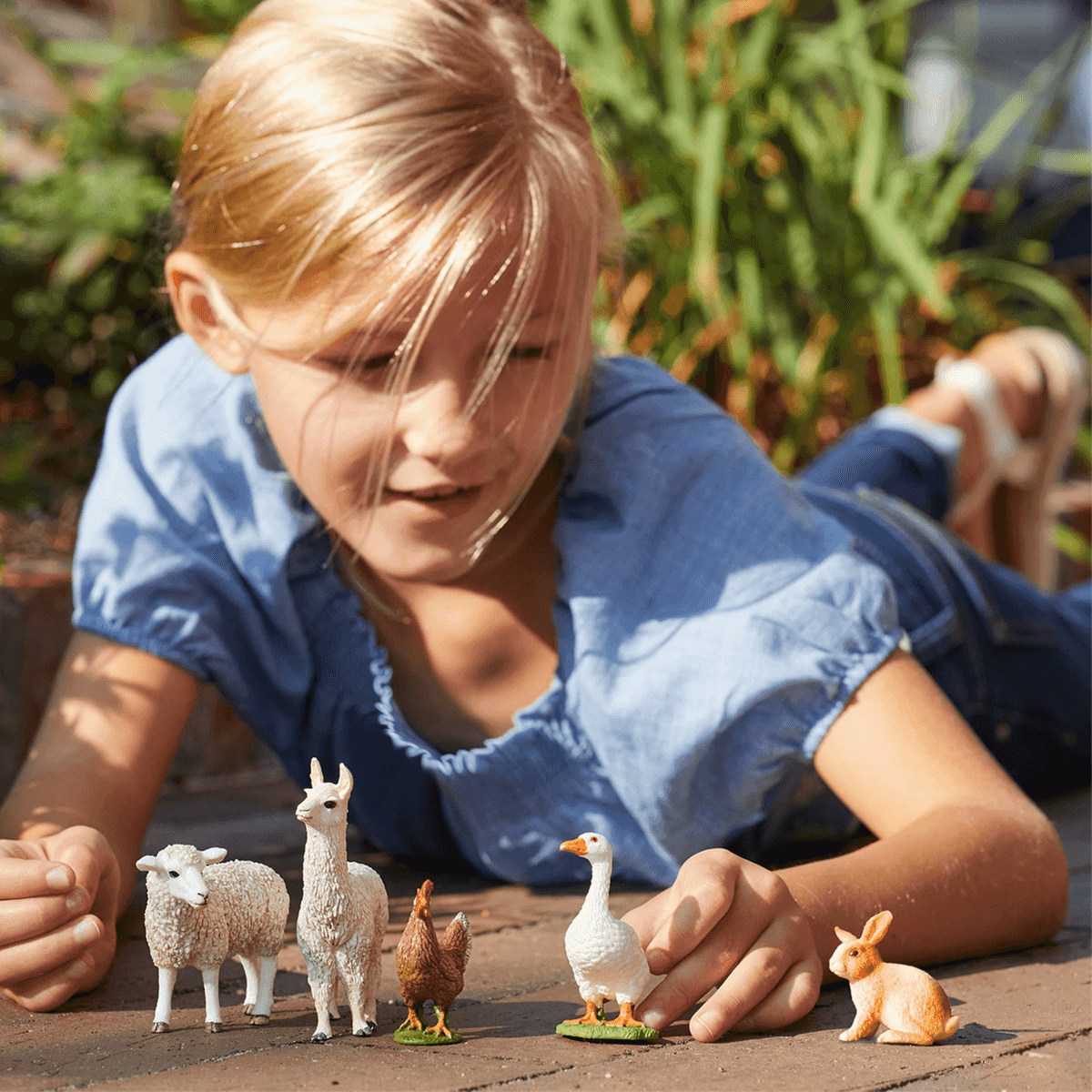 Ein blondes Mädchen liegt auf dem Boden und spielt mit kleinen Tierfiguren wie Schaf, Llama, Huhn, Gans und Hase.