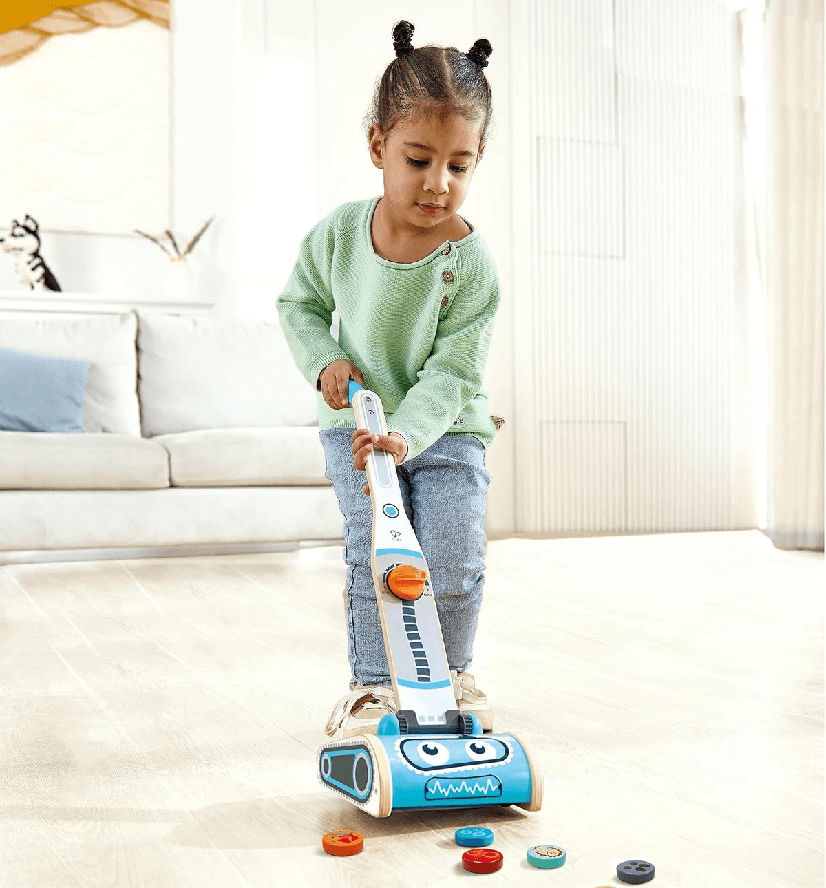 Una niña pequeña con dos rodetes juega con una aspiradora de juguete azul en un suelo de madera.