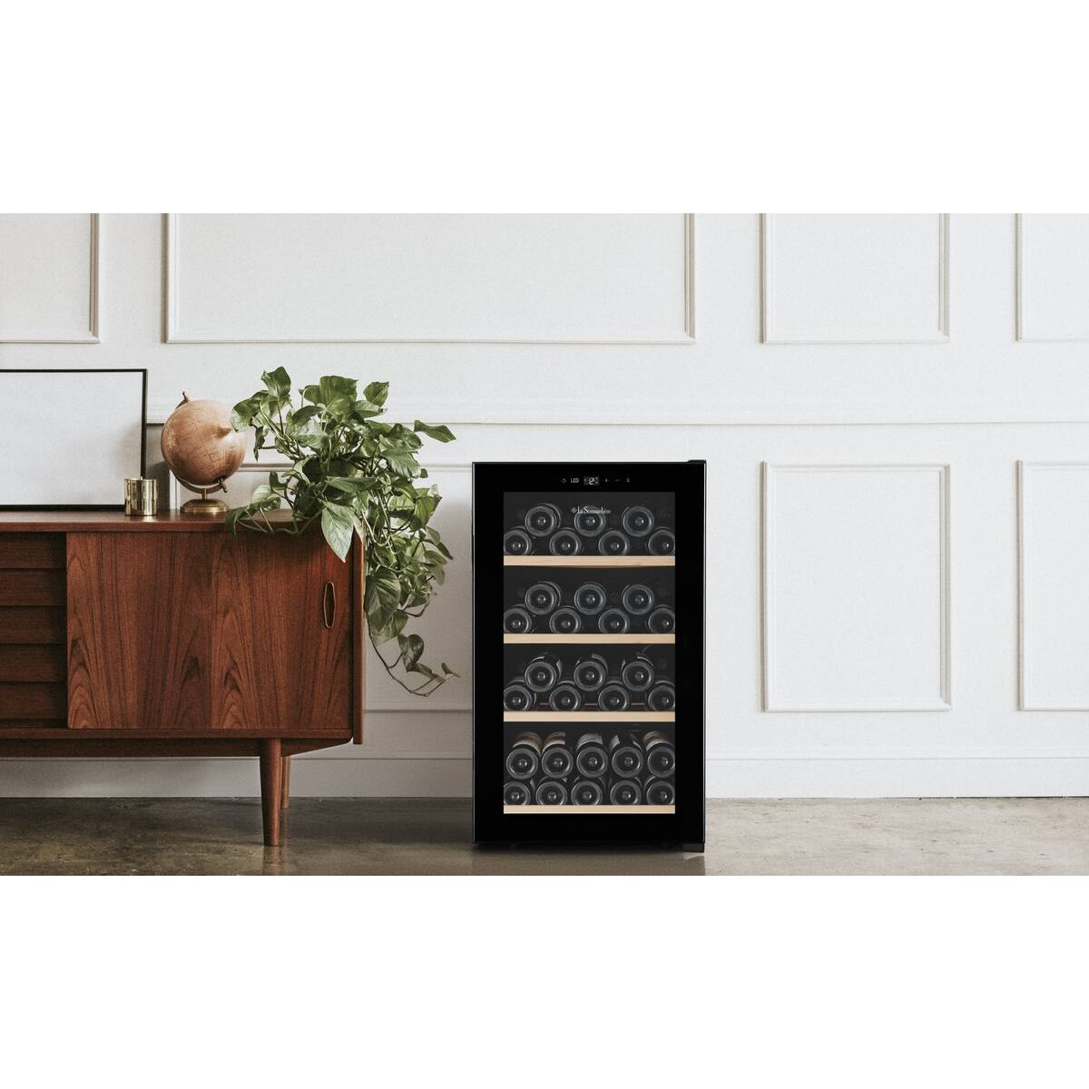 Un enfriador de vino negro se encuentra en una habitación junto a un mueble de madera con plantas.