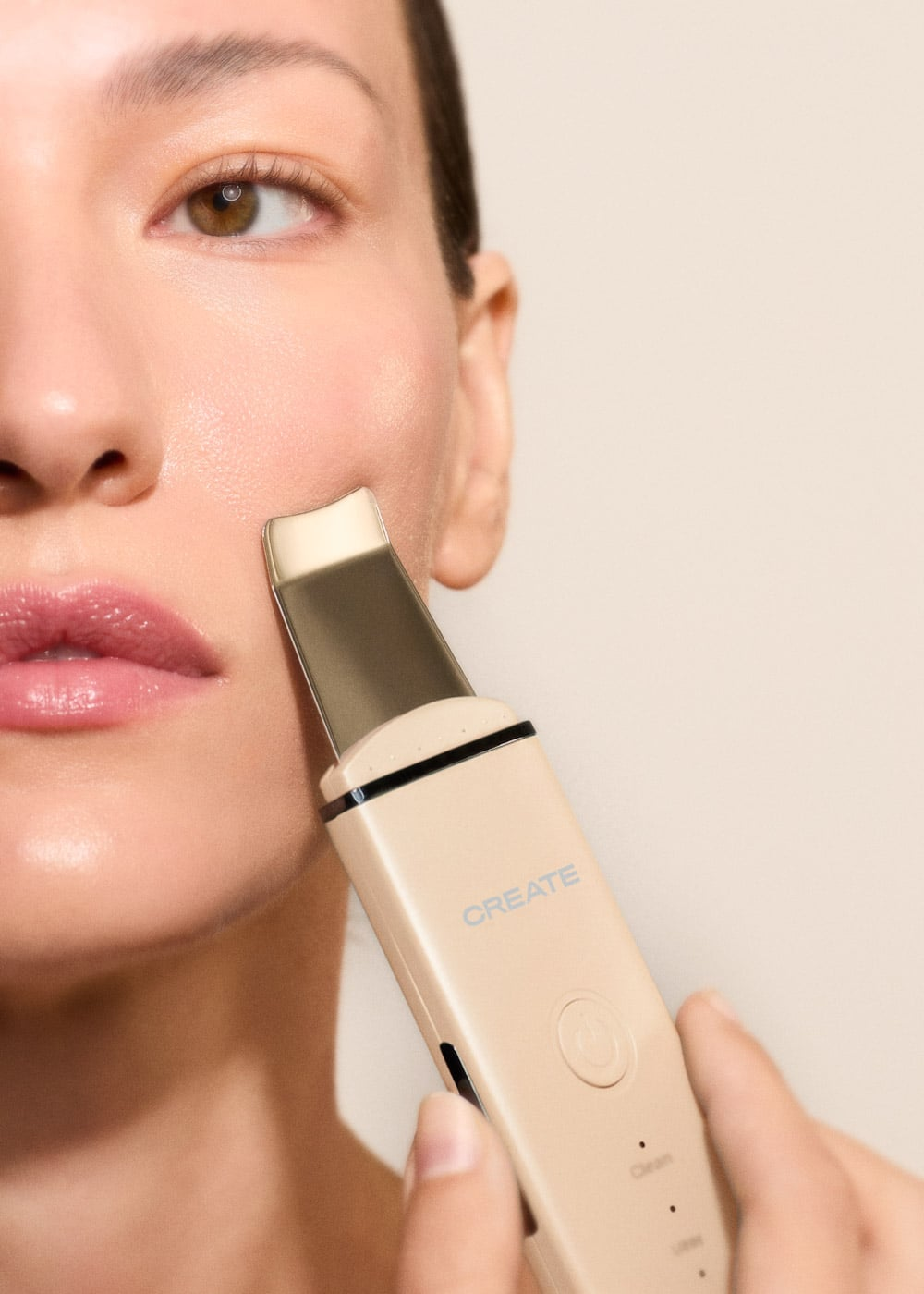 Una mano sostiene una espátula facial ultrasónica beige contra la mejilla de una mujer.