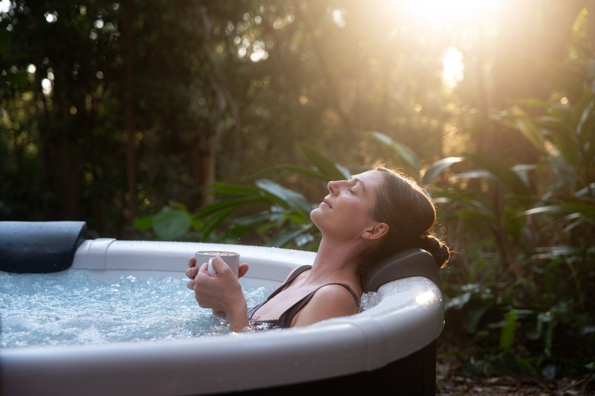 Una mujer se relaja en un jacuzzi al aire libre sosteniendo una taza mientras la luz del sol se filtra entre los árboles.