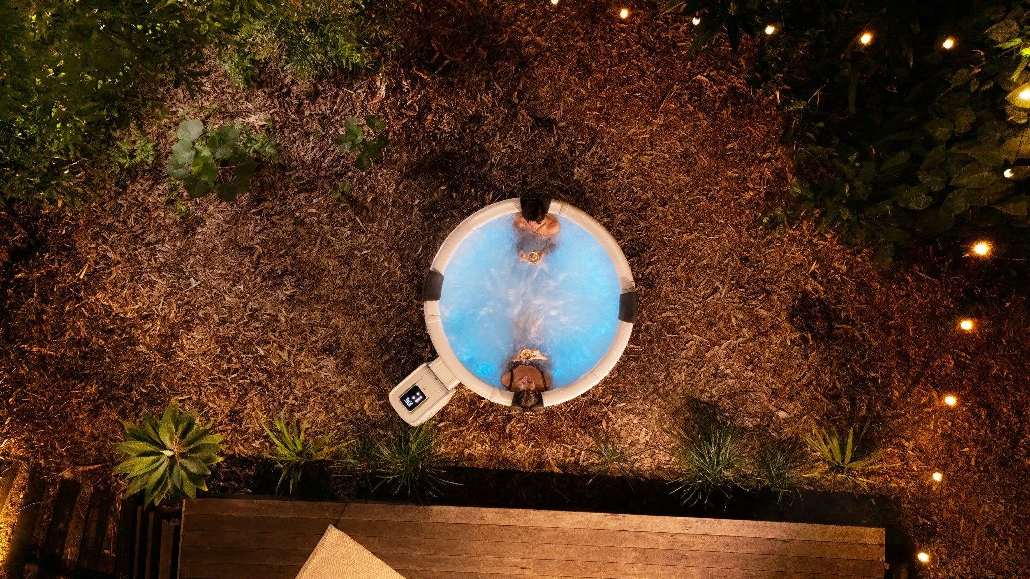 Vista aérea de dos personas relajándose en un jacuzzi iluminado en un jardín por la noche.
