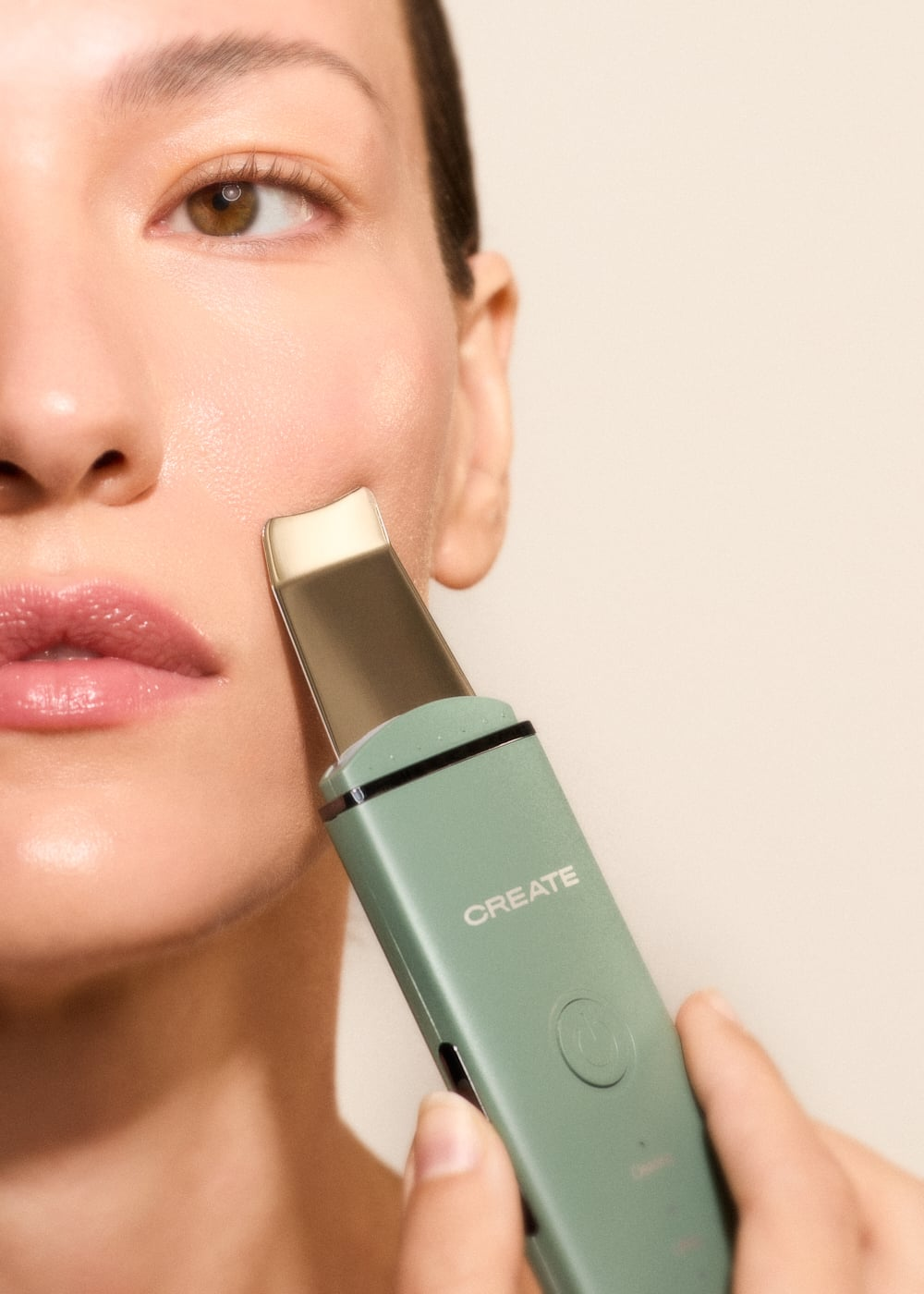 Una mujer usa una espátula facial ultrasónica verde claro en su mejilla.