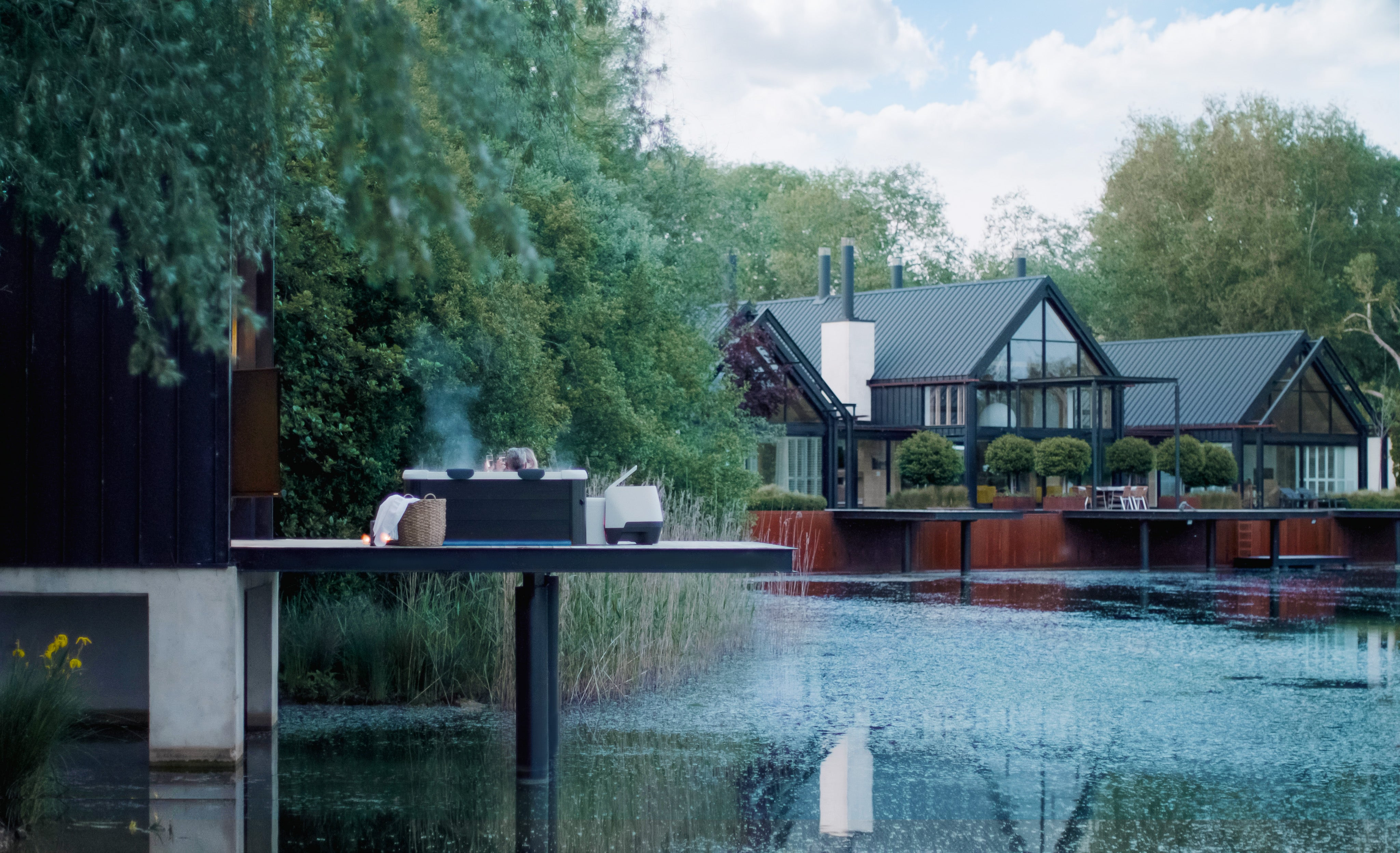Un jacuzzi humeante en una terraza de madera sobre un estanque, con edificios modernos y árboles.