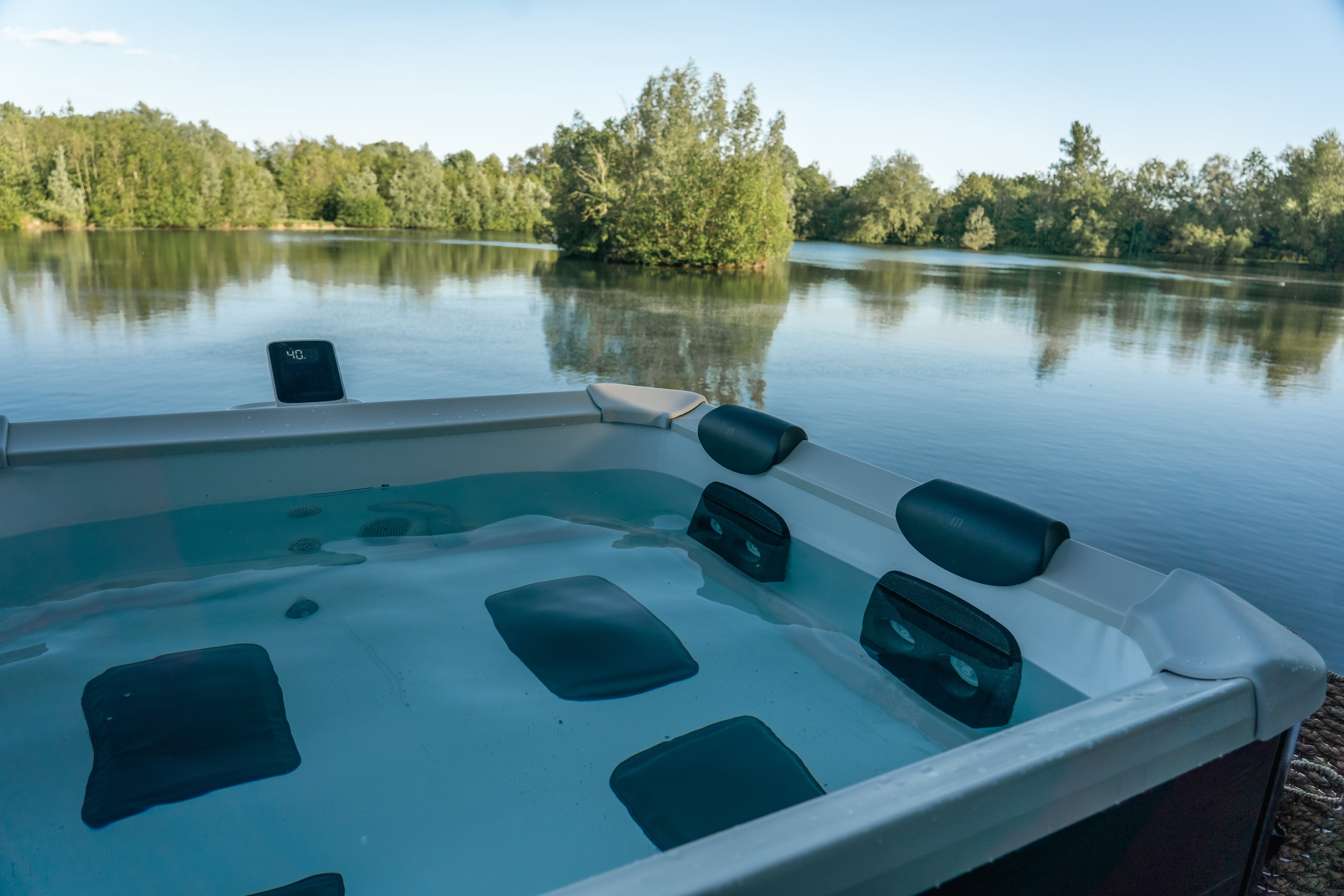 Un jacuzzi al aire libre con agua clara y reposacabezas domina un lago tranquilo rodeado de bosques.