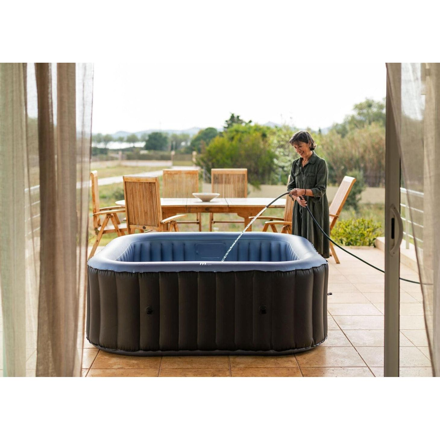 Una mujer llena un jacuzzi inflable con una manguera en una terraza exterior.