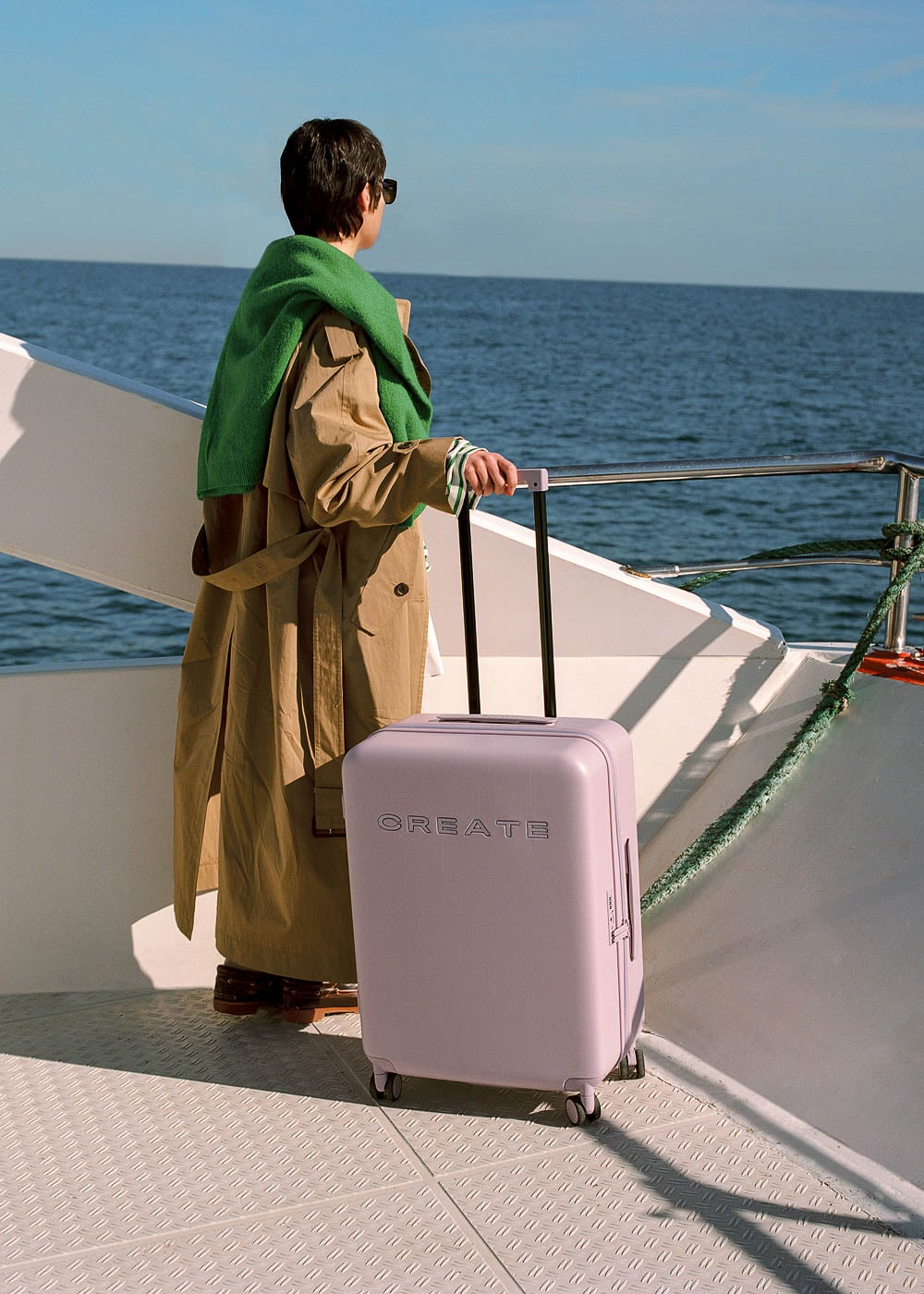 Una persona con gabardina y suéter verde está en un barco junto a una maleta lavanda frente al mar.