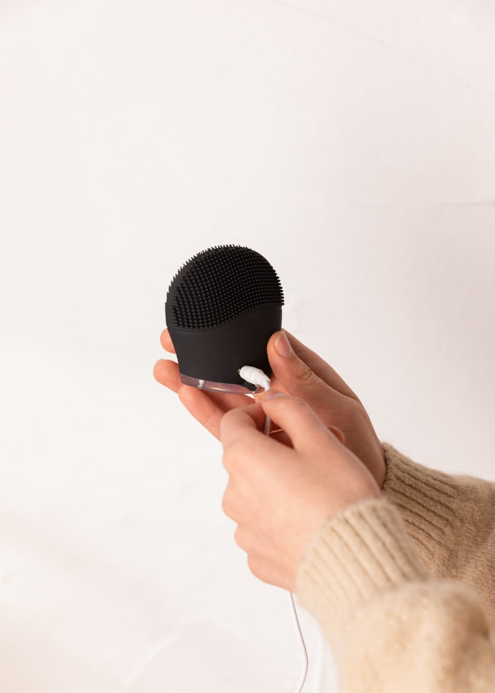 Persona con jersey beis conecta un cable de carga blanco a un cepillo limpiador facial de silicona negro.
