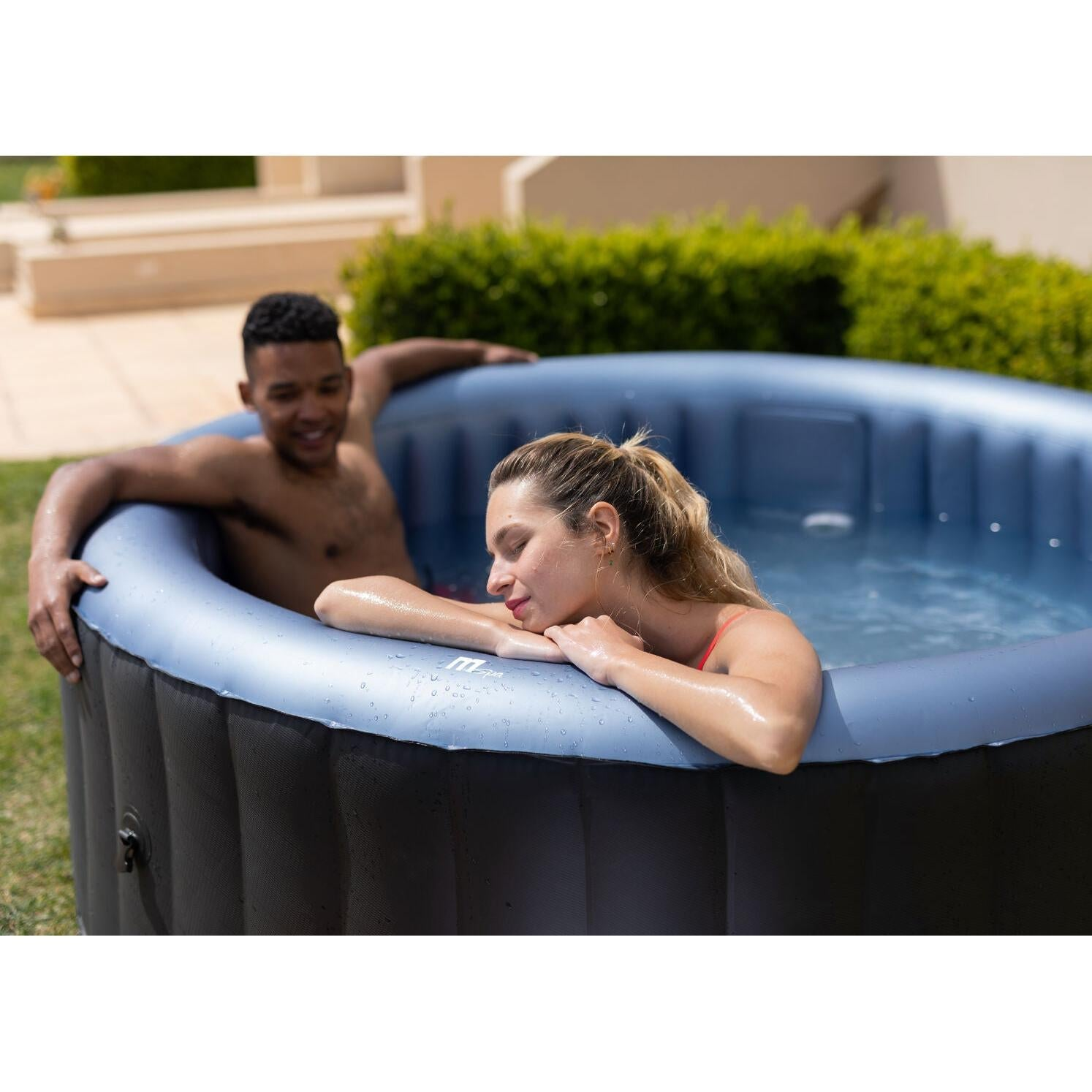 Una pareja se relaja en un jacuzzi inflable al aire libre rodeado de arbustos verdes.