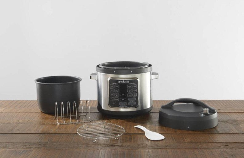 Un multicocina Crockpot plateado con accesorios que incluyen olla interior, rejillas y cuchara sobre una mesa de madera.