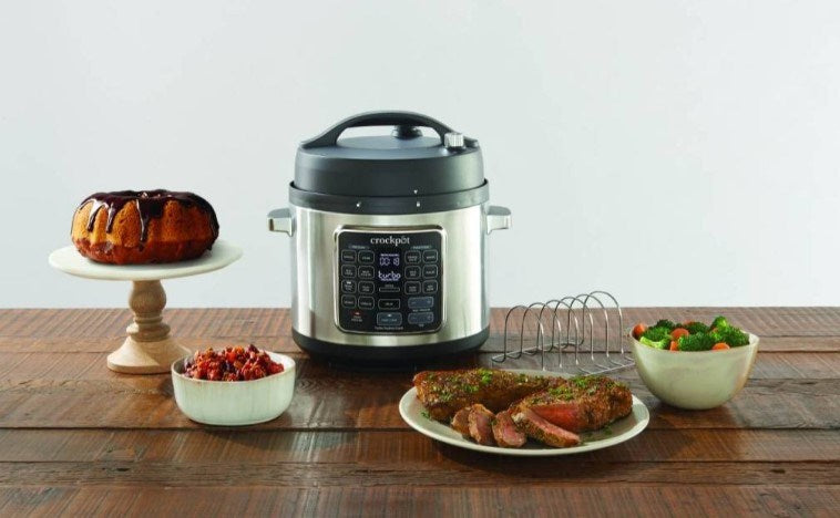 Una olla a presión Crockpot plateada sobre una mesa de madera, rodeada de pan horneado, carne y verduras.