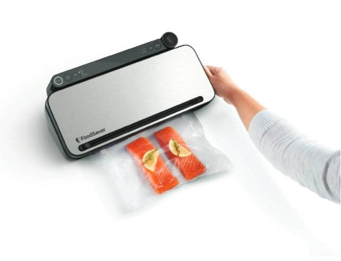 Ręka zamyka srebrną pakowarkę próżniową FoodSaver z dwoma filetami z łososia w plastikowej torbie.