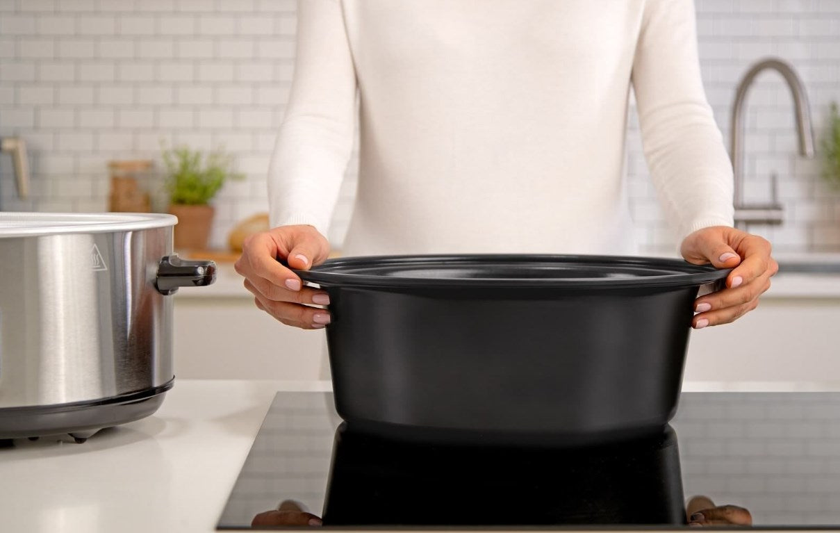 Una persona con suéter blanco levanta una olla de cerámica negra de una placa de cocina con azulejos blancos.