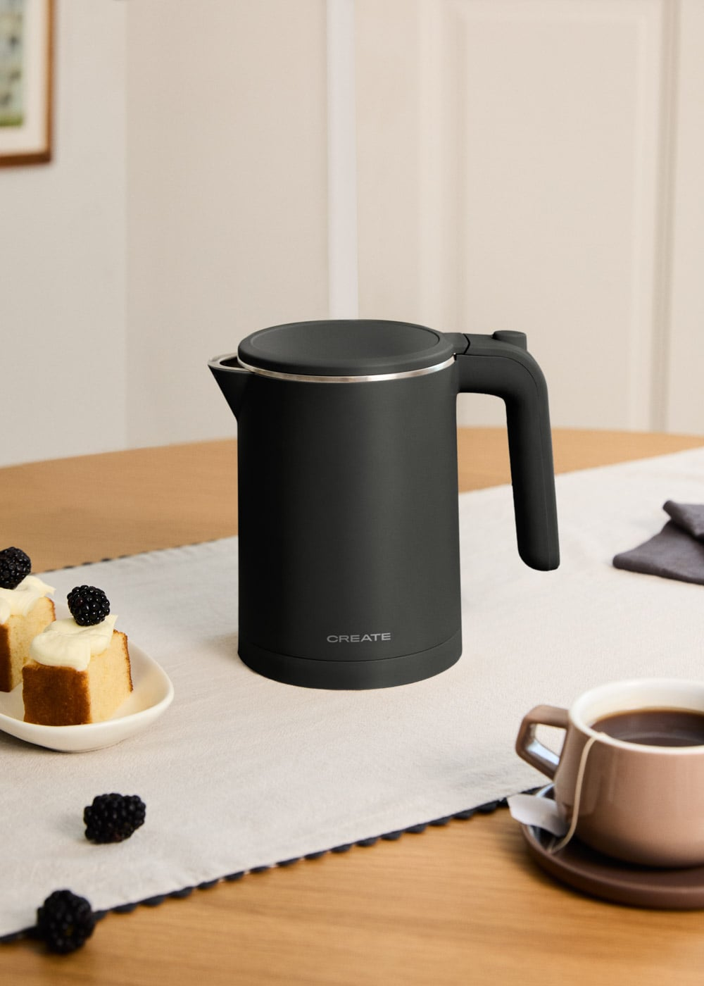 Un hervidor eléctrico negro sobre una mesa con pastel y café.