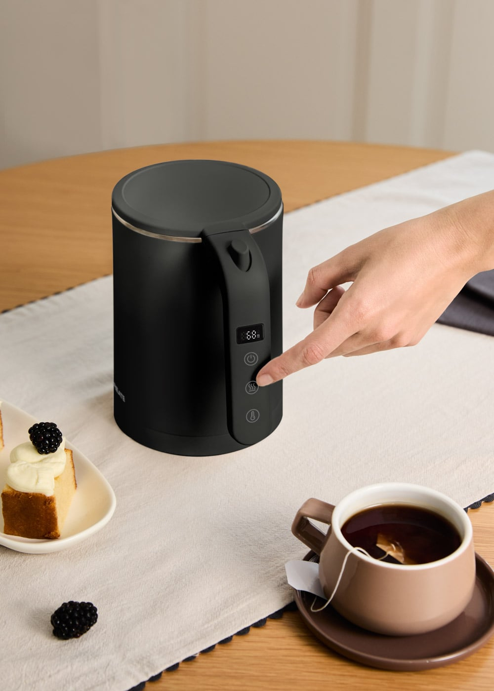 Una mano opera un hervidor eléctrico negro en una mesa con pastel y té.