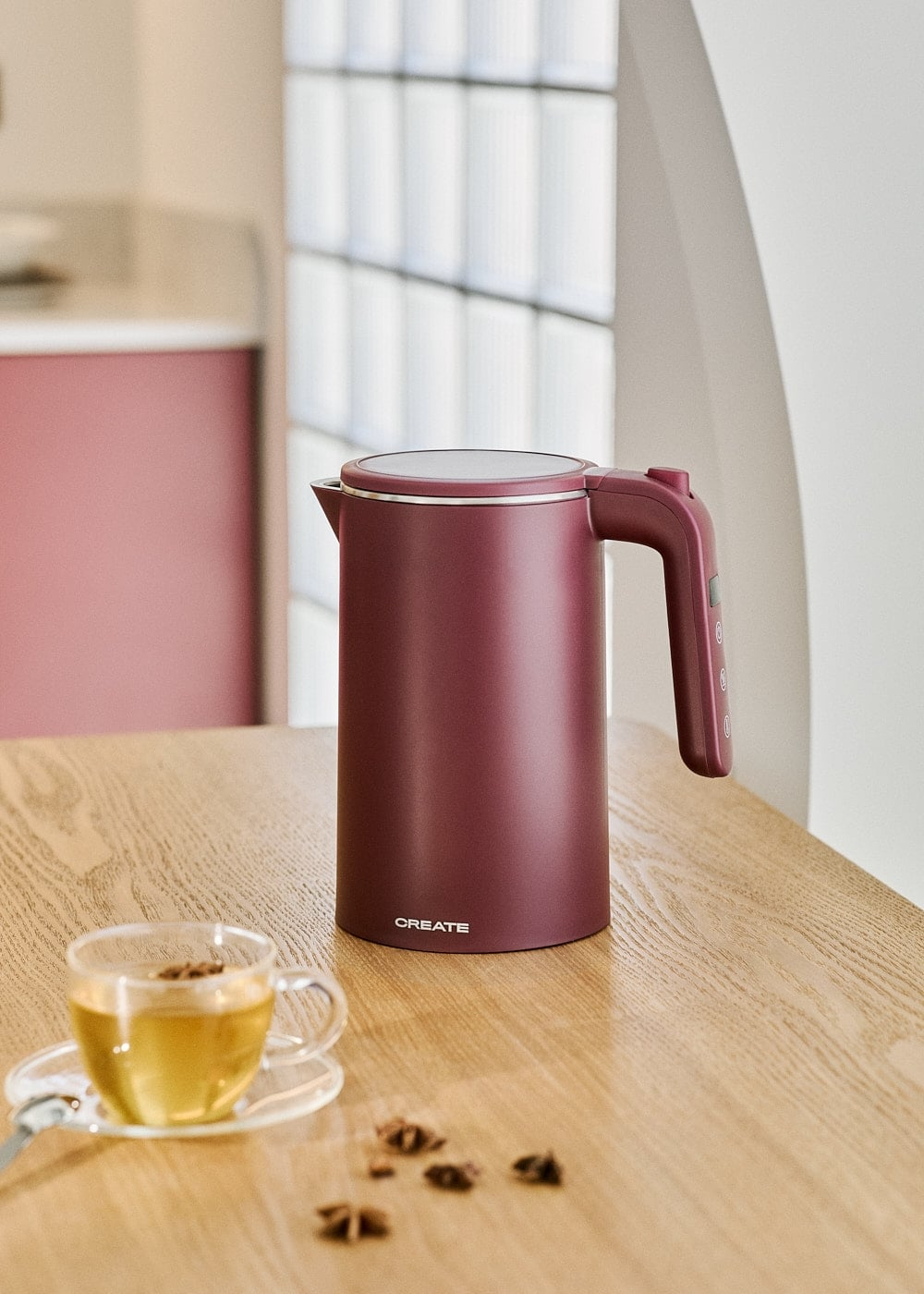 Un hervidor eléctrico granate sobre una mesa de madera junto a una taza de té.