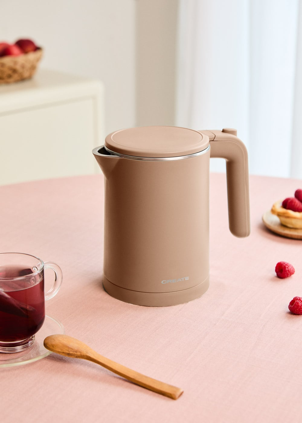 Un hervidor eléctrico beige está sobre una mesa rosa junto a una taza de té y frambuesas.