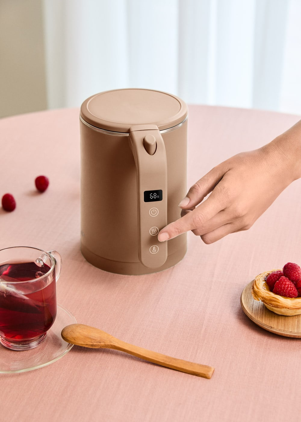 Una mano opera un hervidor eléctrico beige moderno en una mesa rosa con té y repostería.