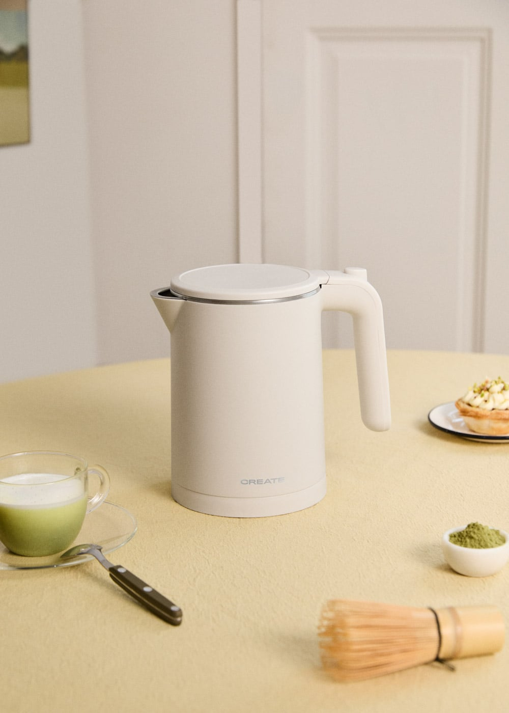 Hervidor eléctrico beis en una mesa con té matcha, pastel y batidor.