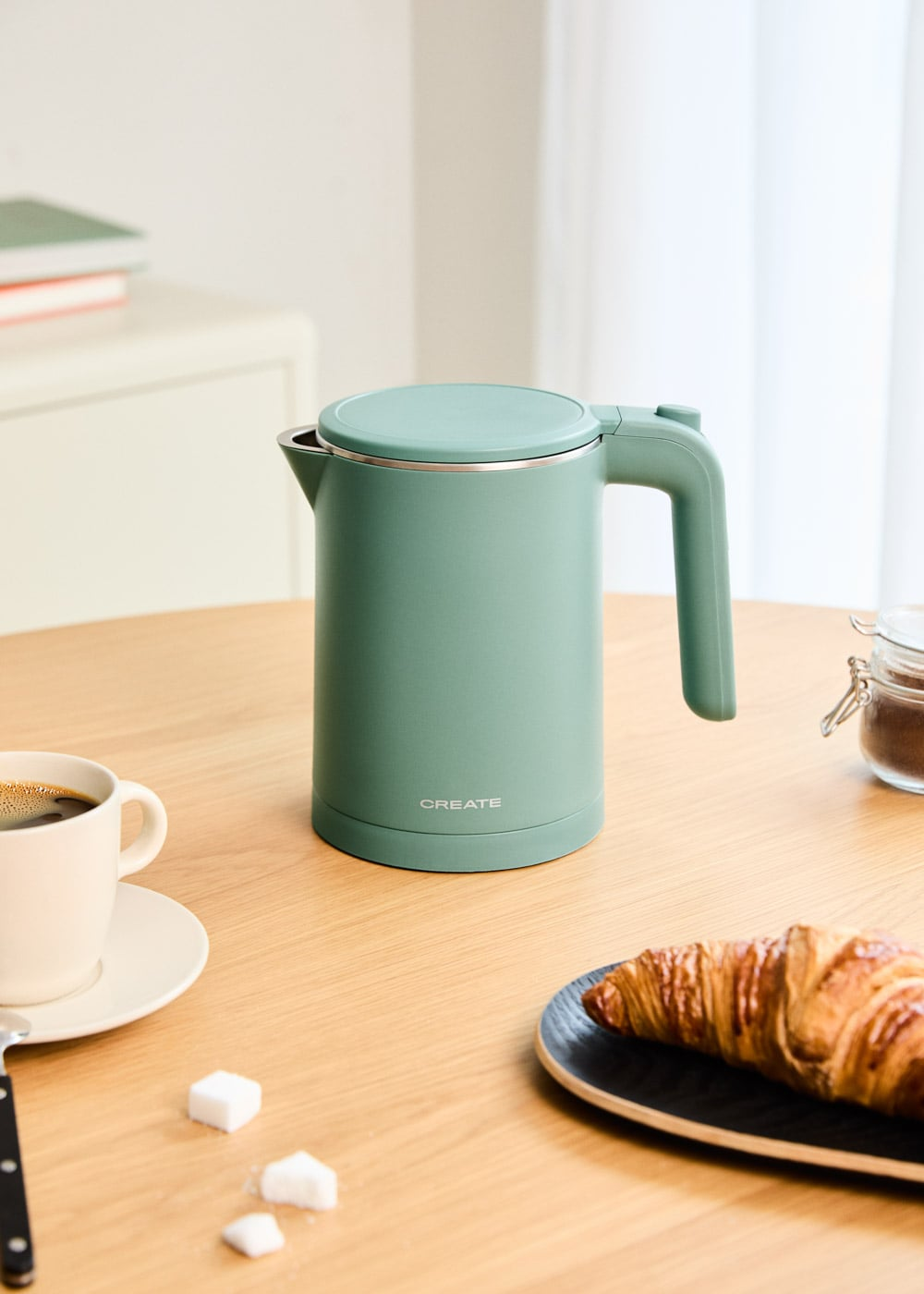 Un hervidor eléctrico verde pastel está en una mesa de madera con una taza de café, un cruasán y terrones de azúcar.