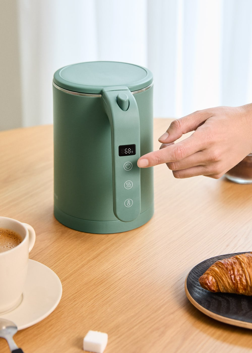 Una mano presiona el botón de encendido de un hervidor verde mate en una mesa de madera con taza y cruasán.