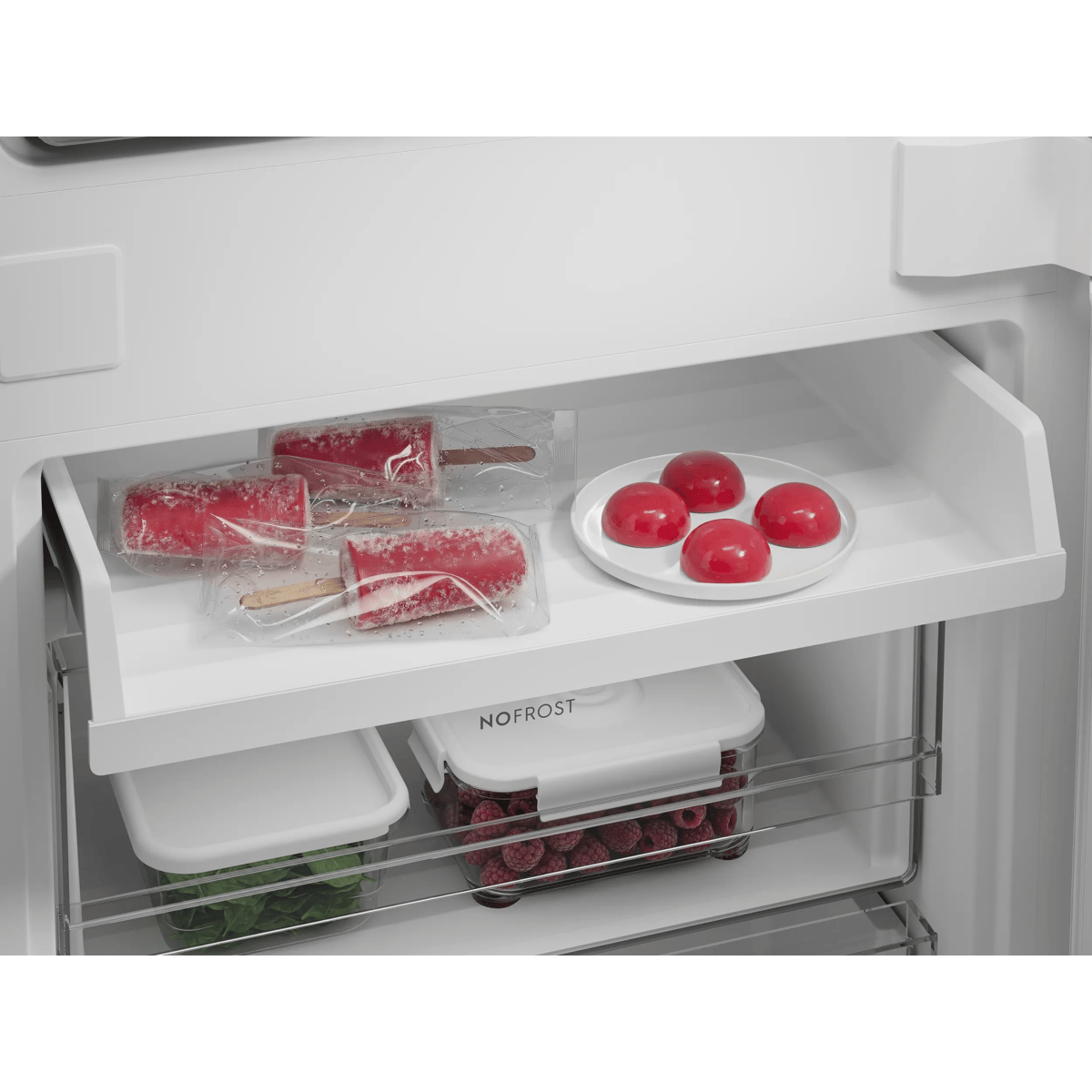 Un congelador blanco con polos envueltos, postres rojos en un plato y recipientes con frambuesas y espinacas.