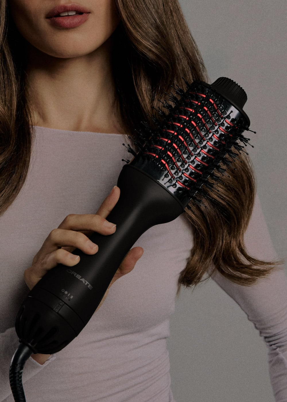 Una mujer sostiene un cepillo moldeador negro y rojo cerca de su cabello castaño.