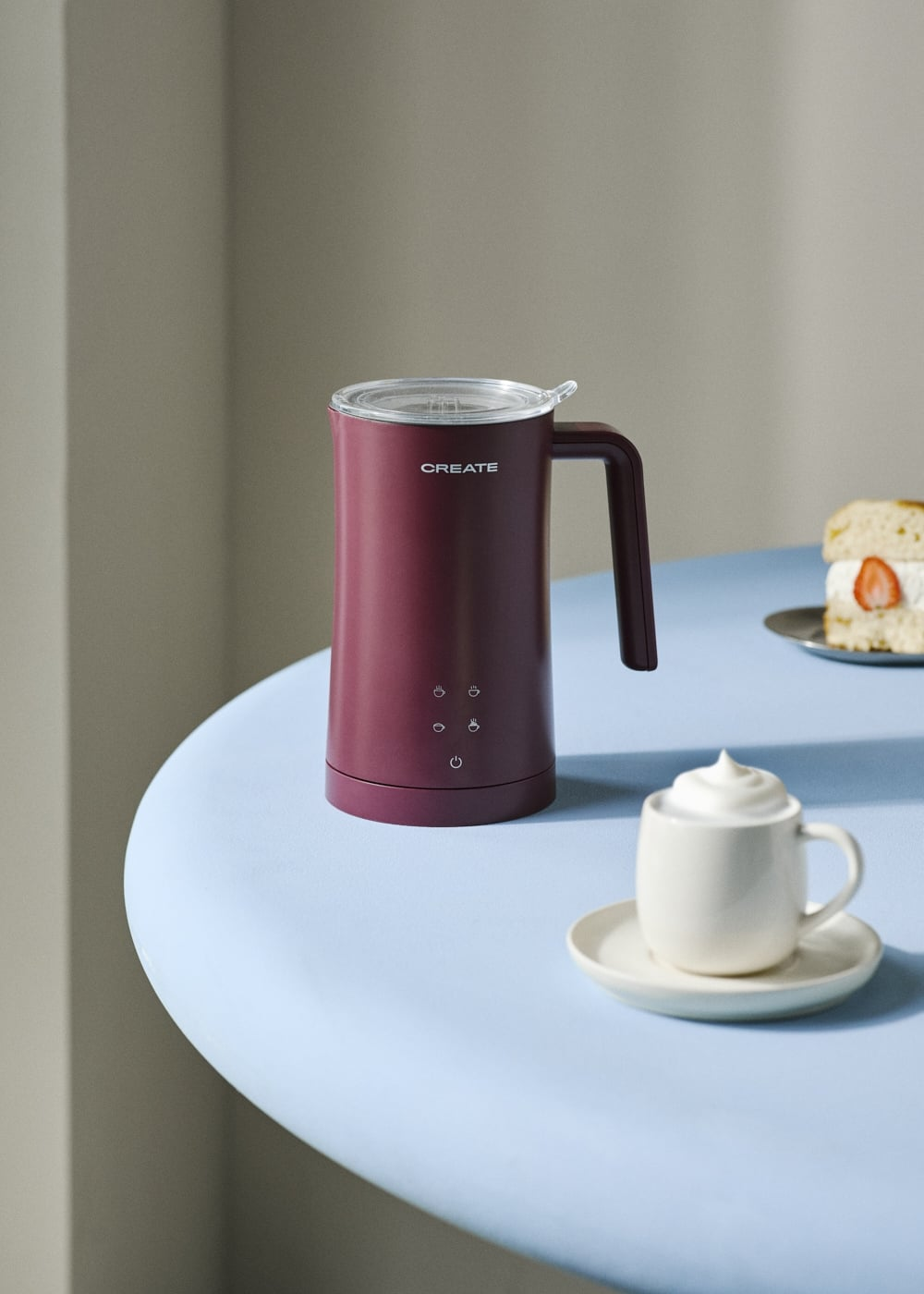 Un espumador de leche burdeos en una mesa azul claro junto a una taza de café.