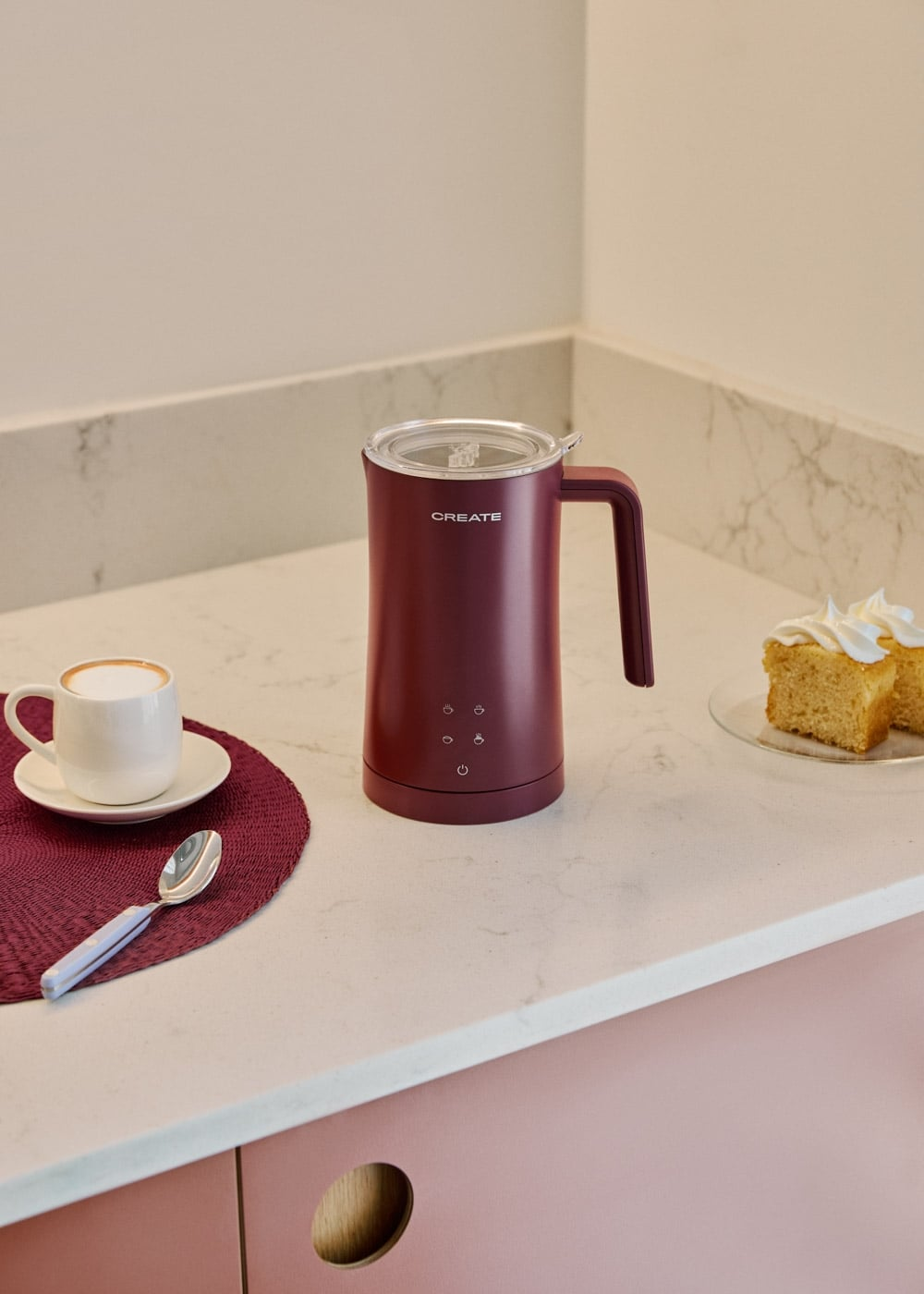 Un espumador de leche granate sobre una encimera blanca con taza, cuchara y pastel.