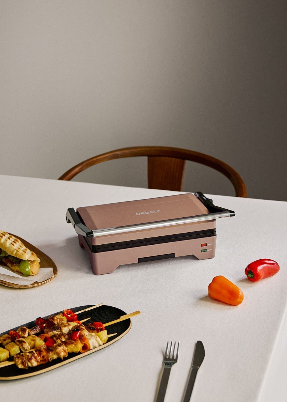 Una prensa para panini rosa en una mesa con brochetas, un sándwich, pimientos y cubiertos.