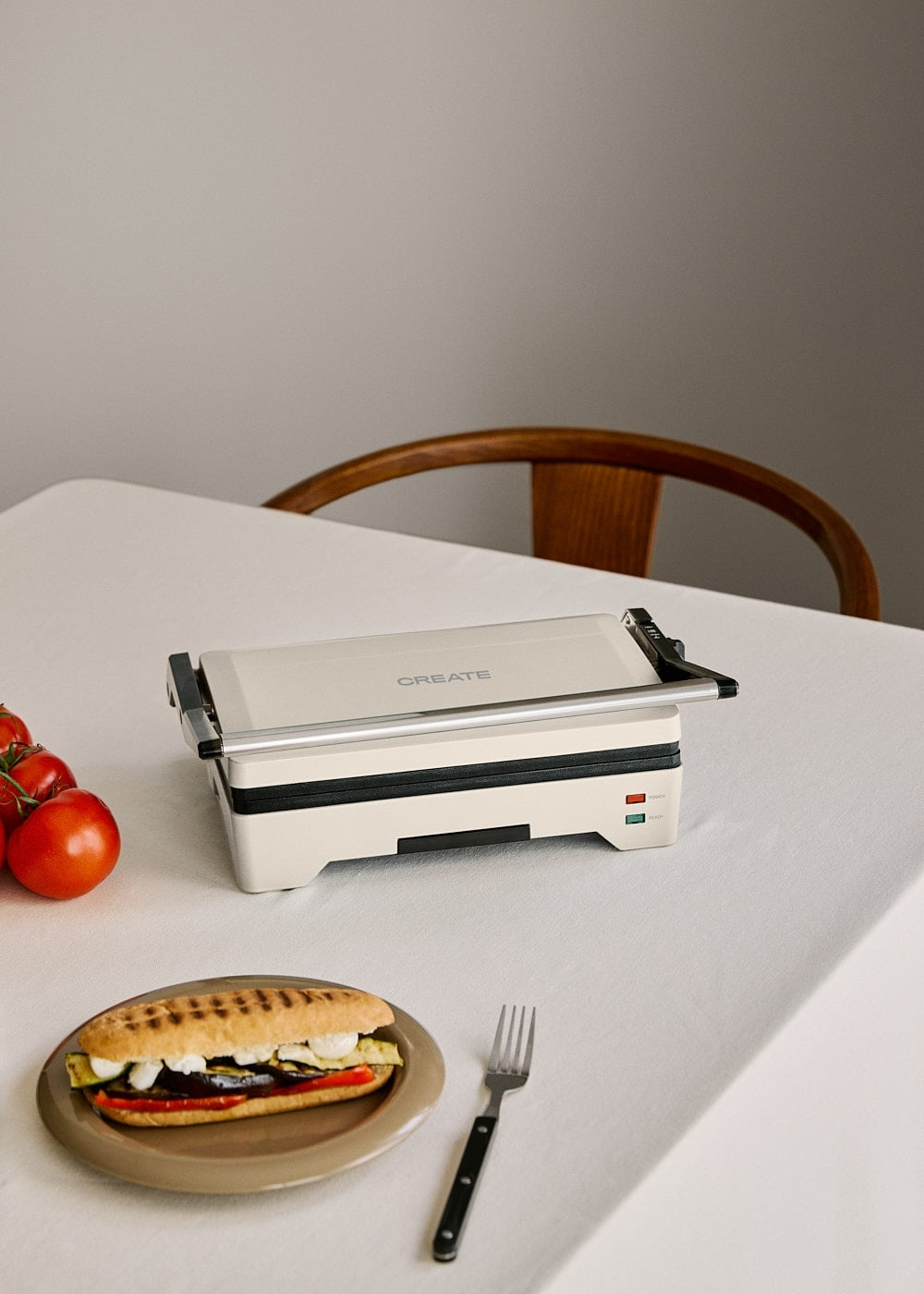 Una tostadora de sándwiches blanca en una mesa, junto a tomates, un panini en un plato y un tenedor.