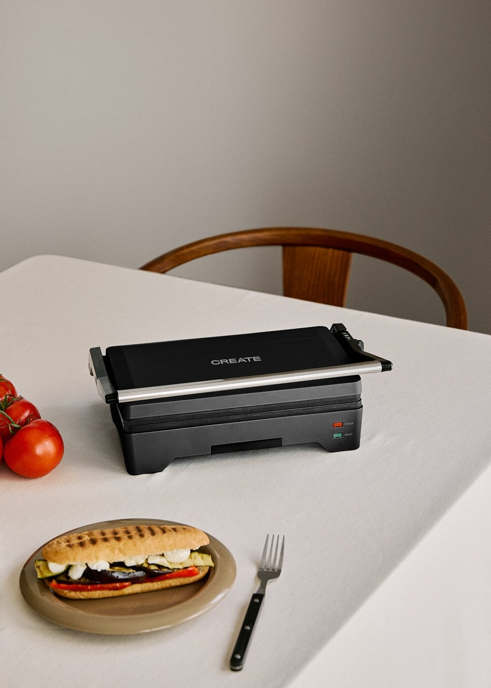 Una parrilla para panini negra se encuentra en una mesa blanca junto a un panini y tomates frescos.