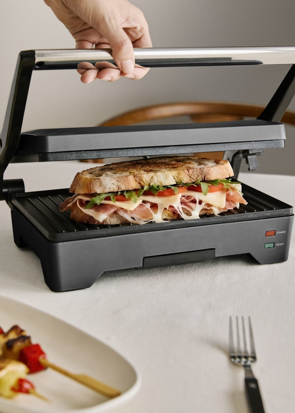 Una mano abre una paniniera negra que revela un sándwich a la parrilla con queso fundido, jamón, tomates y verduras.