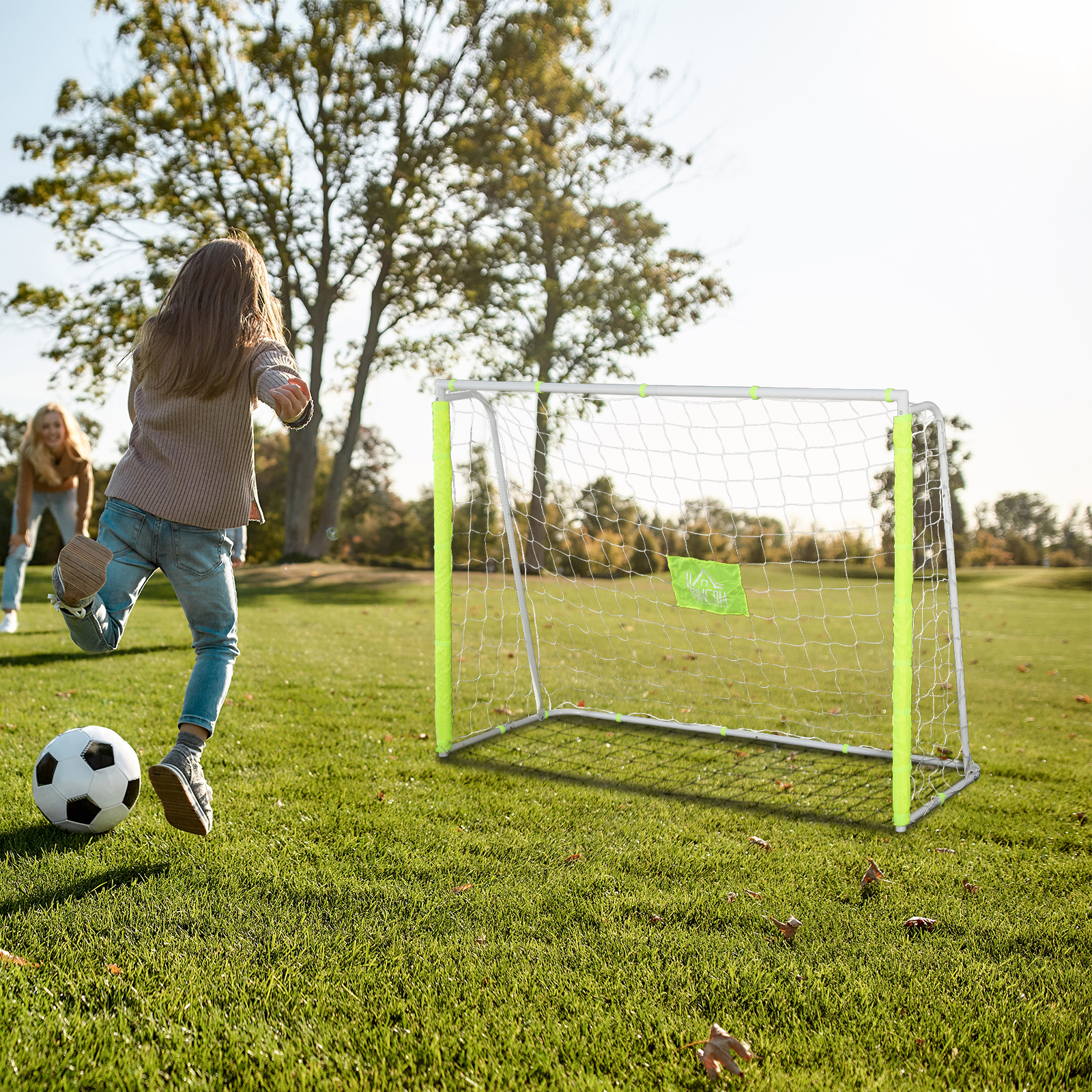 Un niño patea un balón hacia una portería de fútbol en un campo de césped, con otra persona observando.