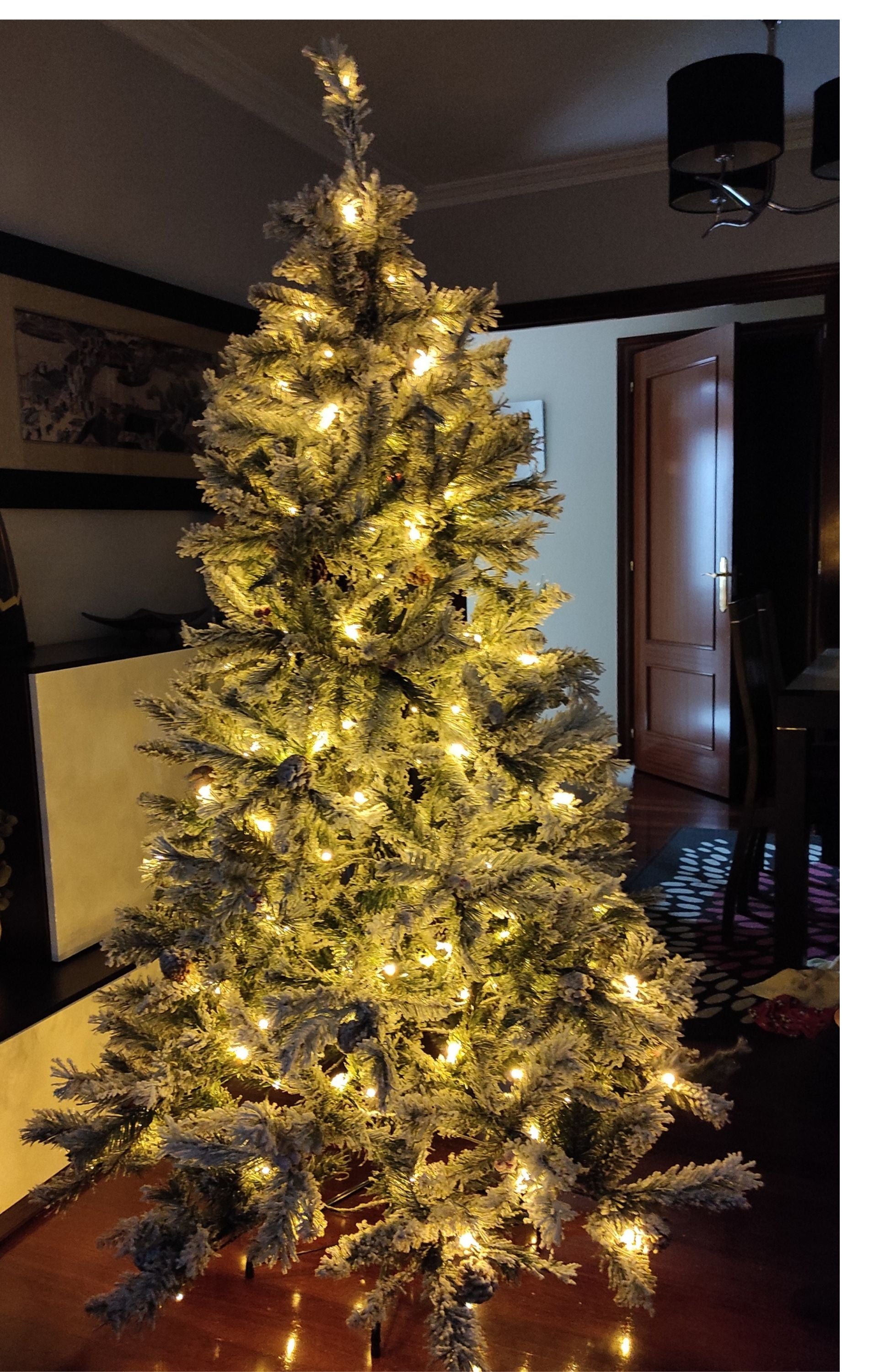 Un árbol de Navidad nevado con luces cálidas se encuentra en una habitación tenuemente iluminada con una puerta abierta.