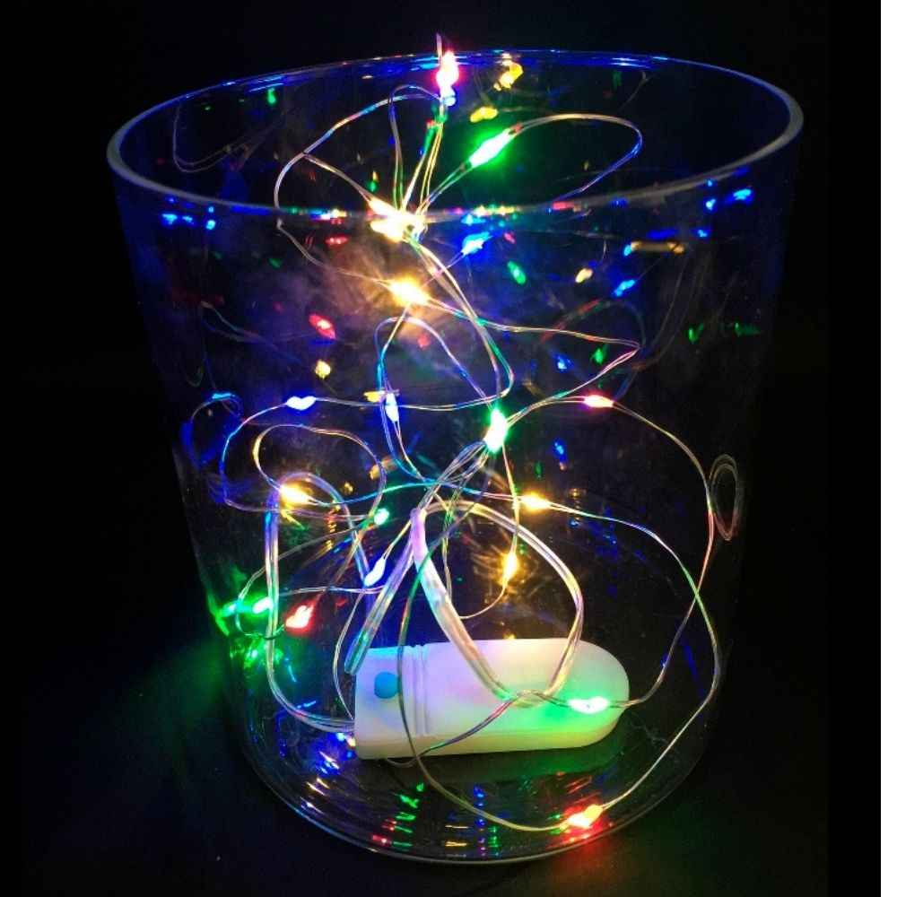 Un frasco de vidrio transparente sobre fondo negro lleno de luces de hadas coloridas y brillantes y una caja de pilas blanca.