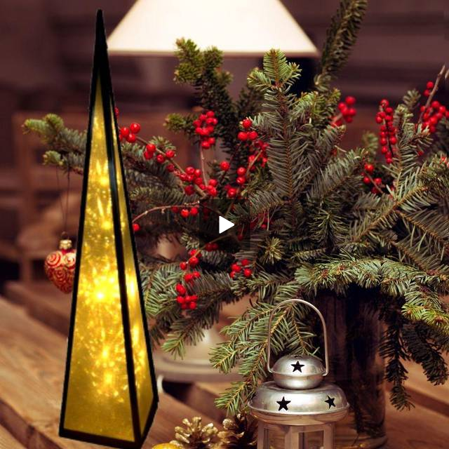 Una lámpara triangular, ramas navideñas con bayas rojas y una linterna plateada en una mesa de madera.
