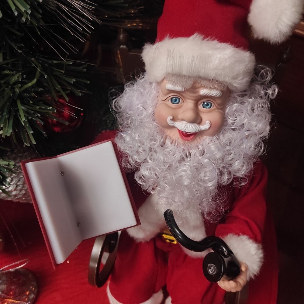 Muñeco de Papá Noel con barba blanca sostiene un libro rojo abierto y una pipa negra frente a un árbol de Navidad.