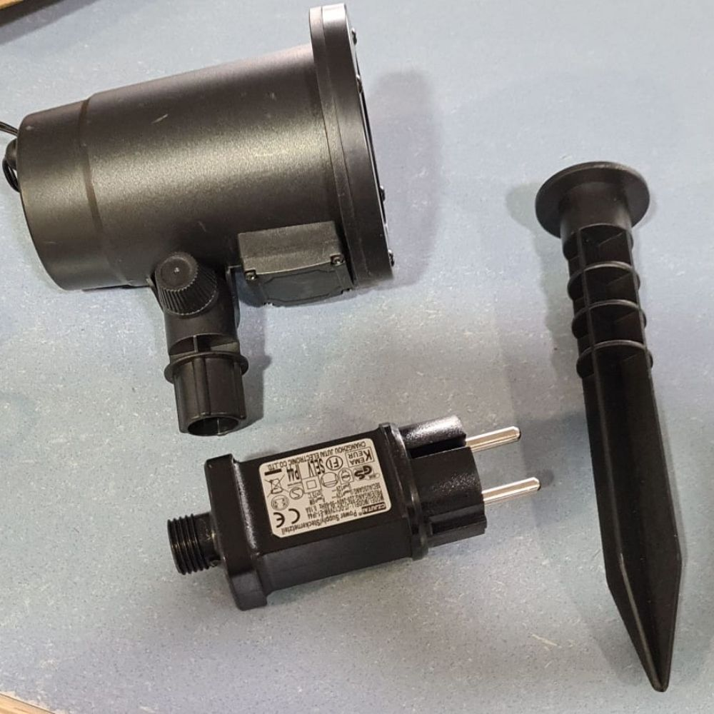 Un foco negro para exteriores, adaptador de corriente y estaca en una superficie gris.