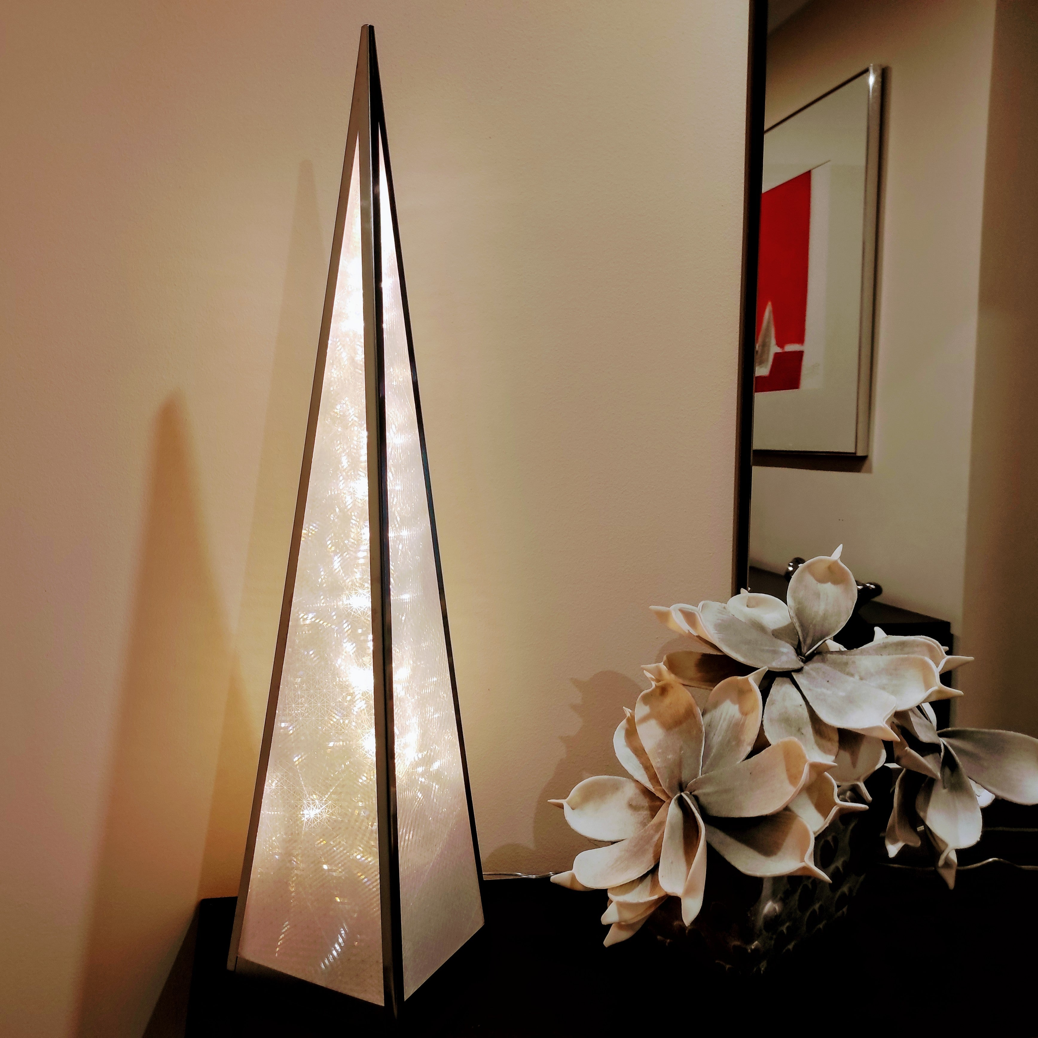 Una lámpara piramidal iluminada y triangular se encuentra junto a unas flores sobre una mesa.