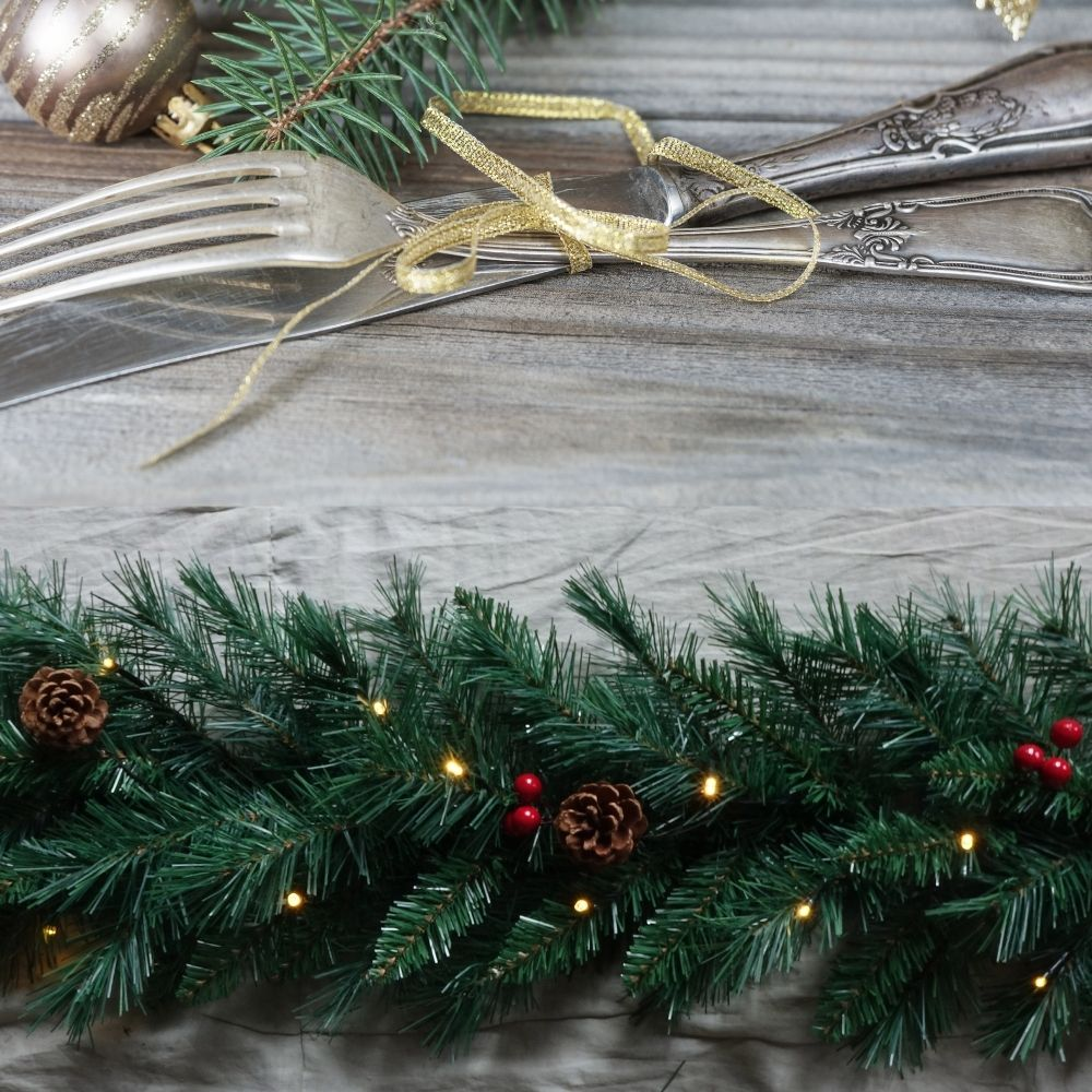 Decoración navideña con cubiertos de plata, ramas de pino y luces en madera.