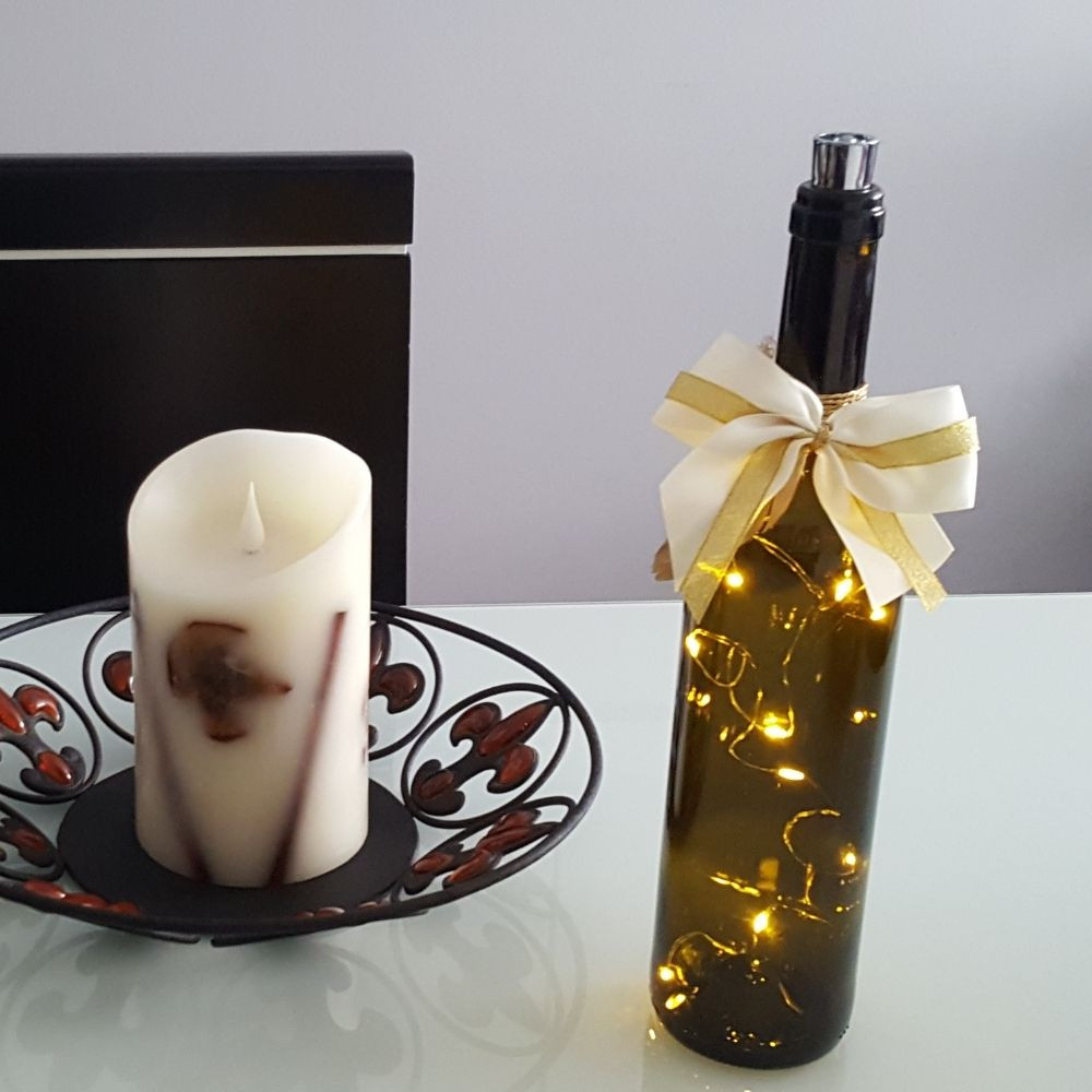 Una vela blanca en un cuenco de metal y una botella de vino con luces y lazo blanco en una mesa.